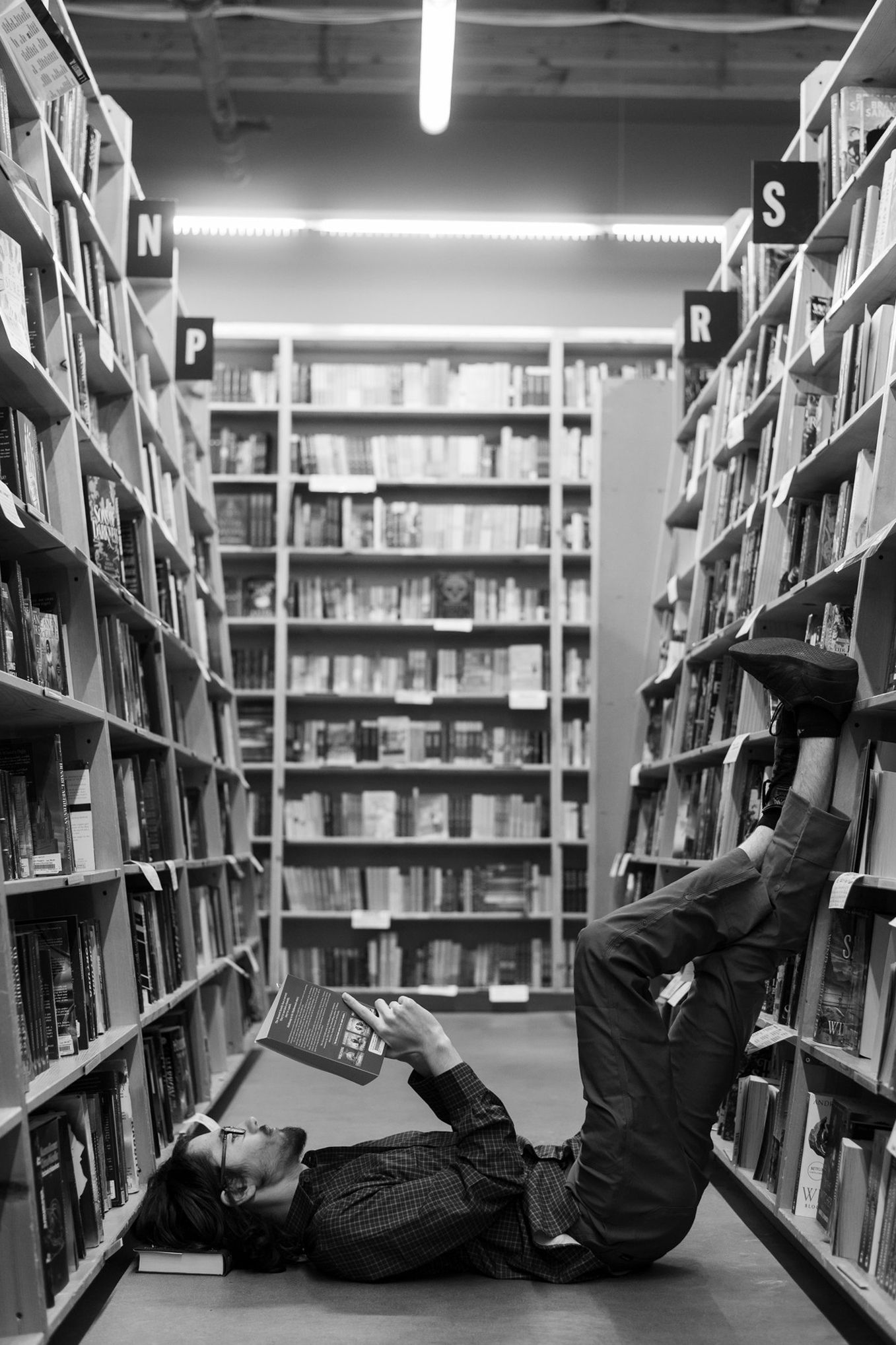 A creative black and white photo of someone lying sideways between library bookshelves while reading.