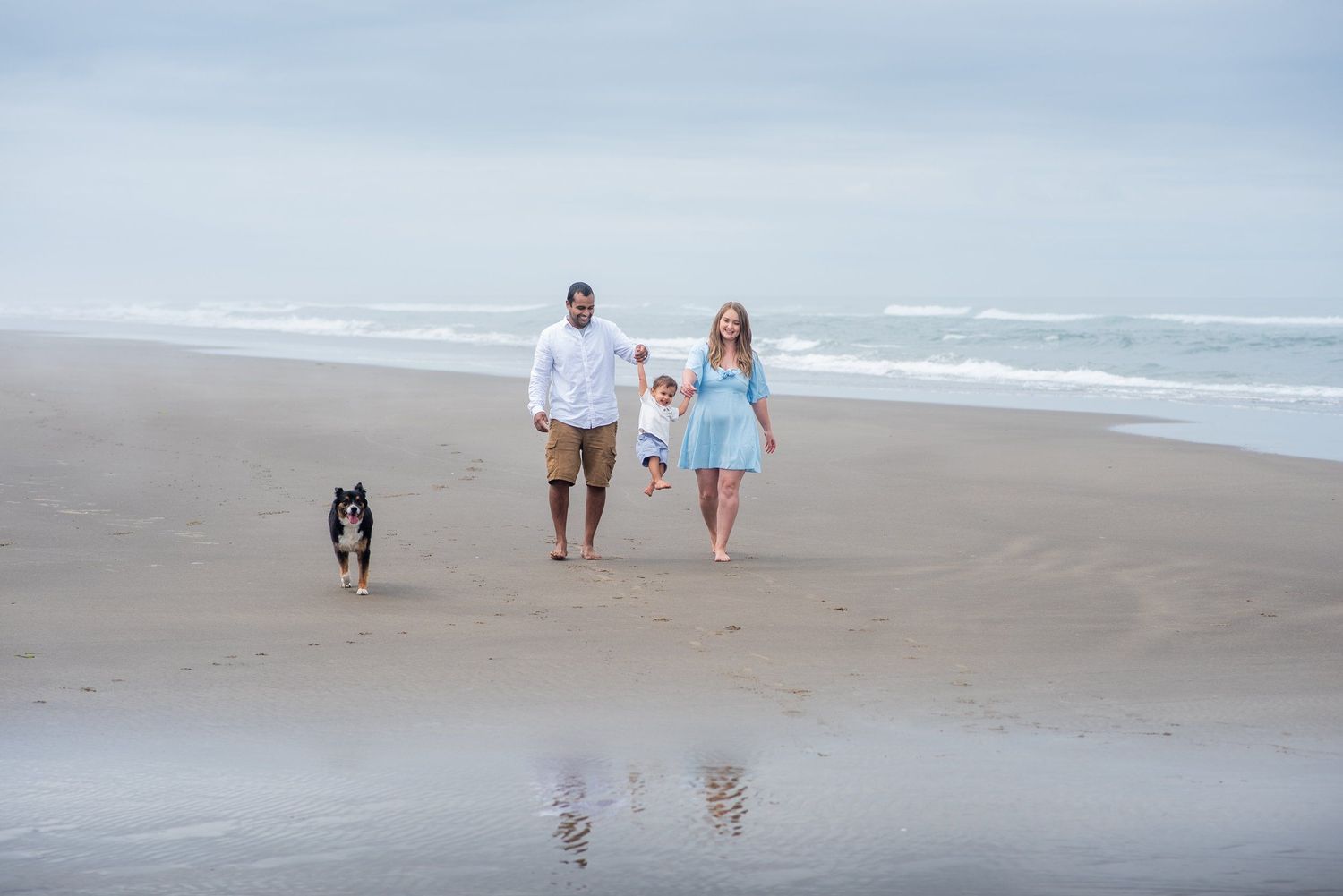 Family walking along peaceful beach shoreline with dog during overcast day.