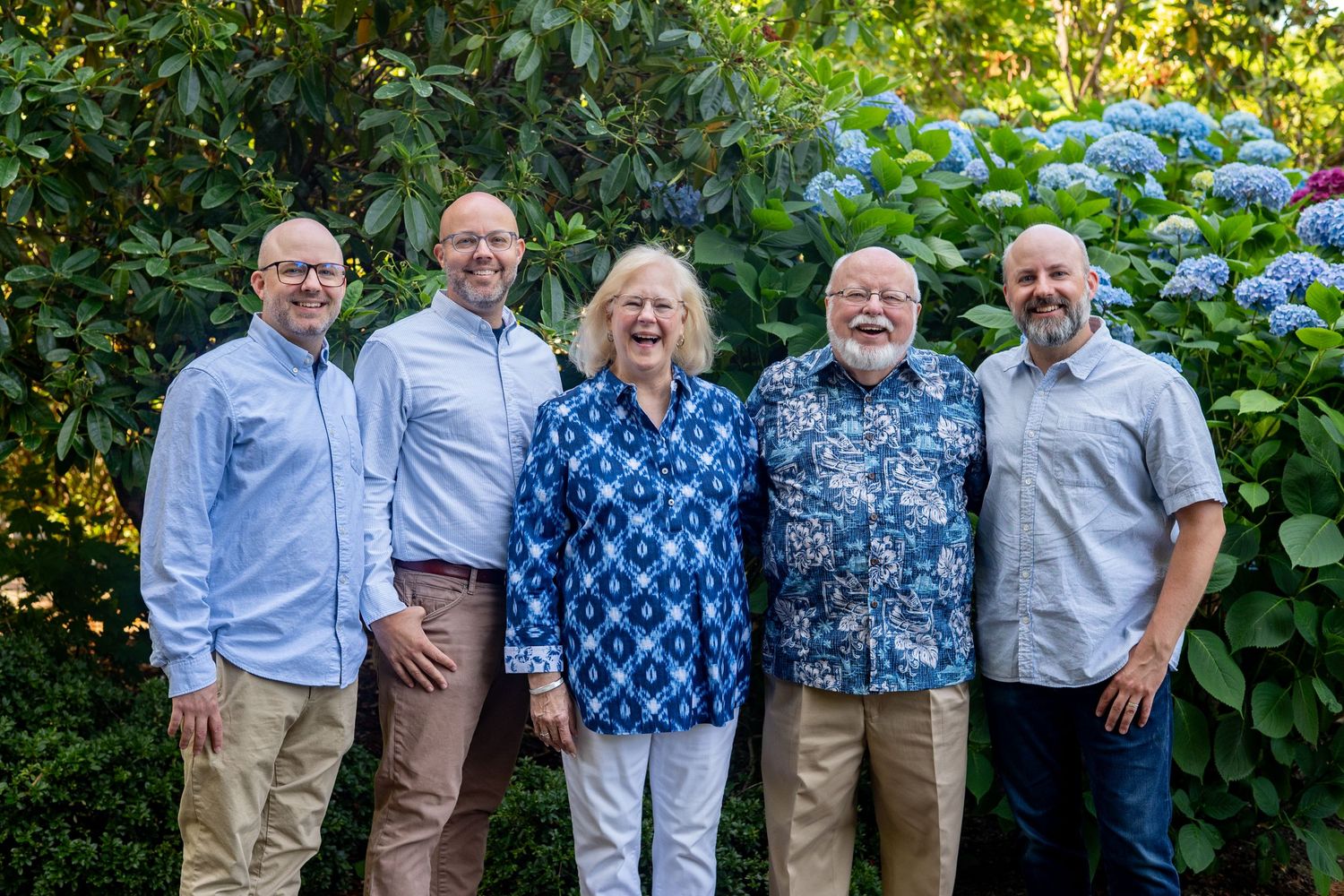 A group of friends stand together smiling in front of blooming hydrangea bushes in a garden setting.