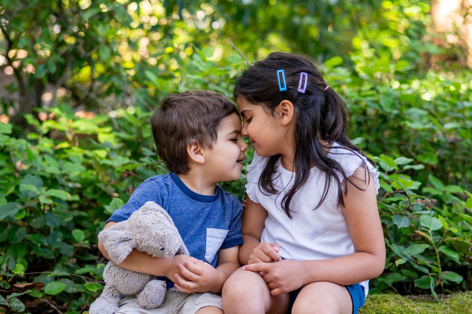 Two young children sit together in a lush green garden having an intimate conversation in the sunlight.