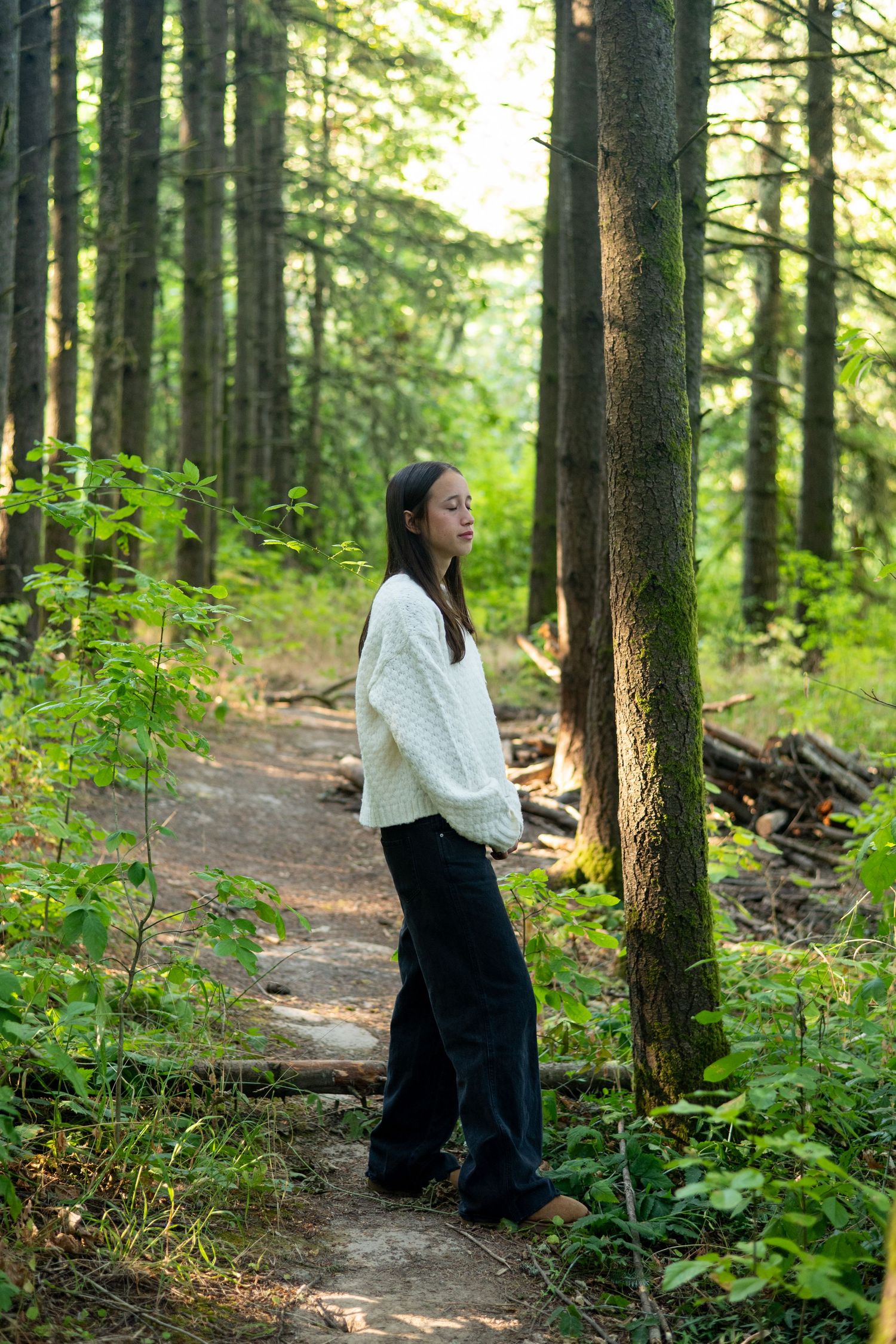 Person in a white knit sweater and dark pants standing on a forest trail surrounded by evergreen trees in soft sunlight.