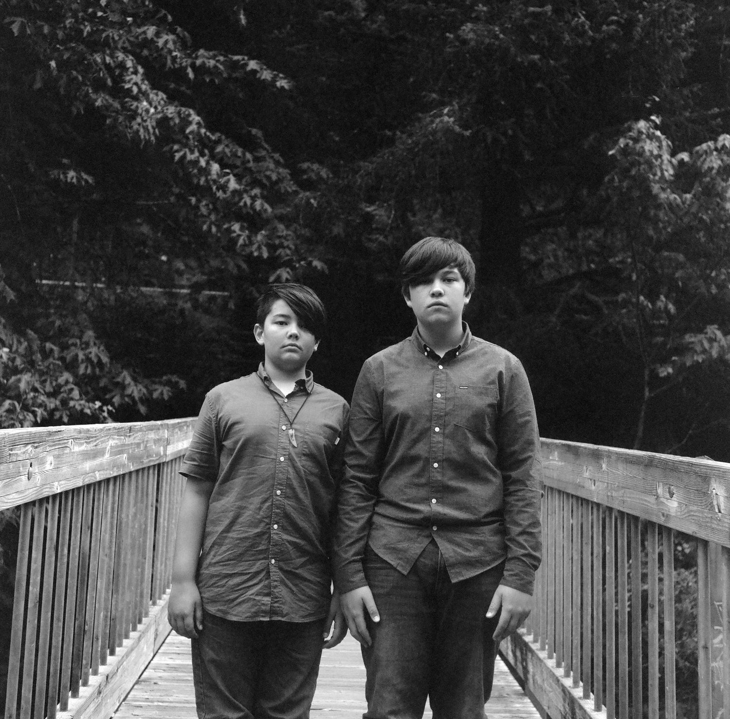 Two people walking along a wooden footbridge surrounded by trees in black and white.