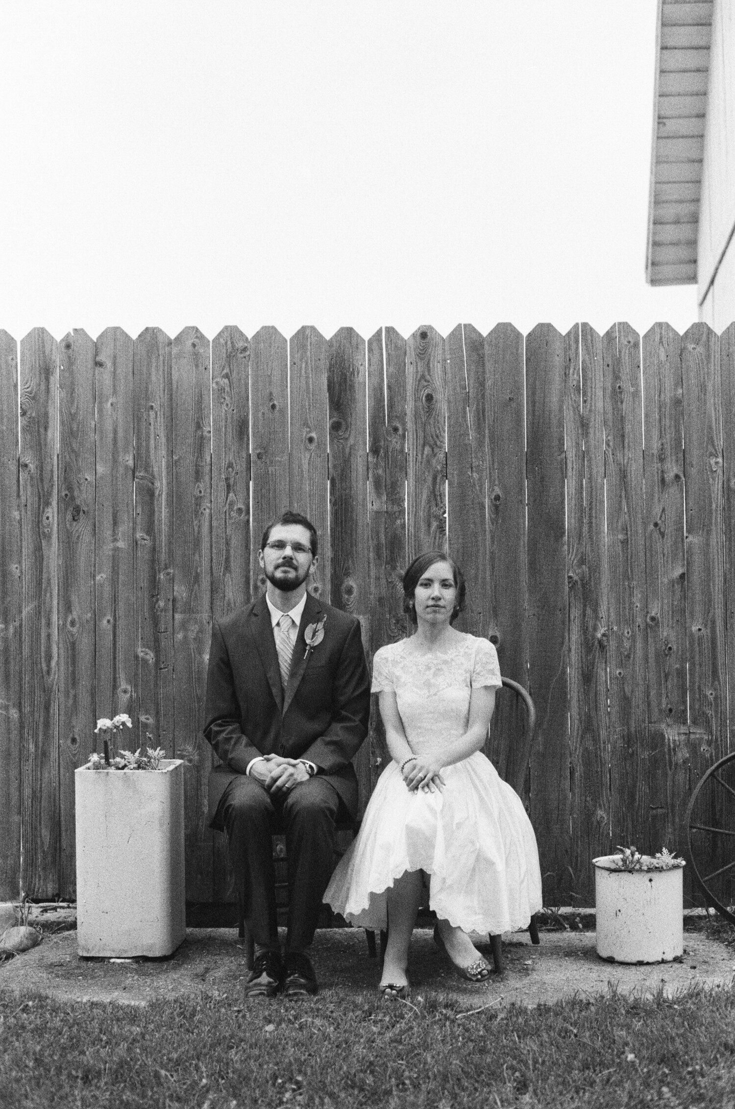A couple in wedding attire sits on a bench against a wooden fence with potted flowers.