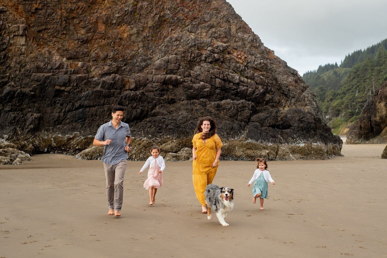 A family running together with their dog on a scenic beach with dramatic rock formations.