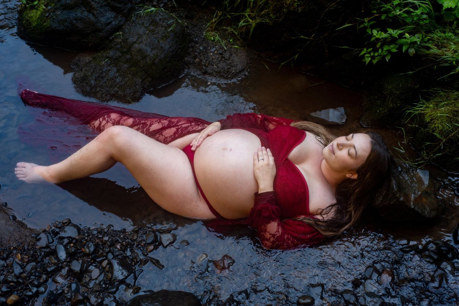 maternity session with woman in red dress reclining on rocks near dark flowing stream in natural outdoor setting.