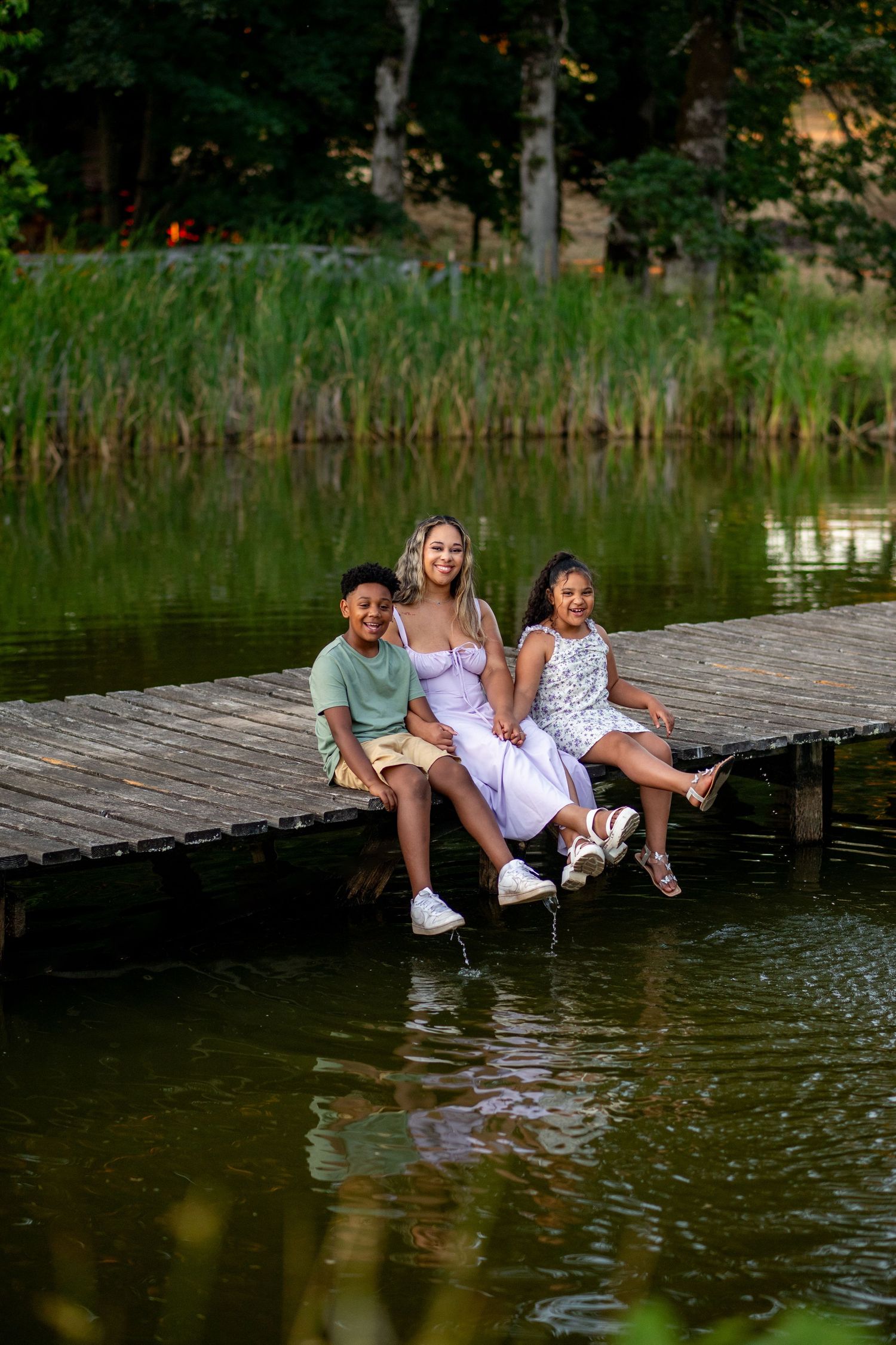 A family sits together dangling their feet off a wooden dock over a tranquil pond on a summer afternoon.