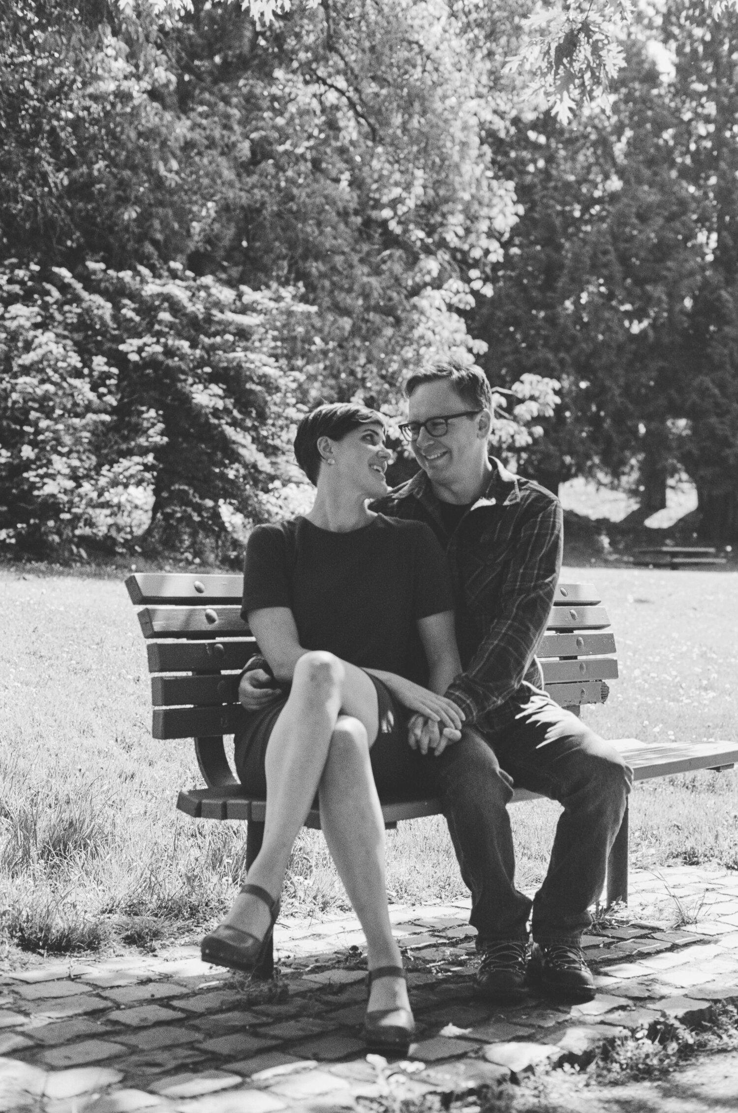 A couple sits together on a wooden park bench surrounded by trees in a black and white photograph.