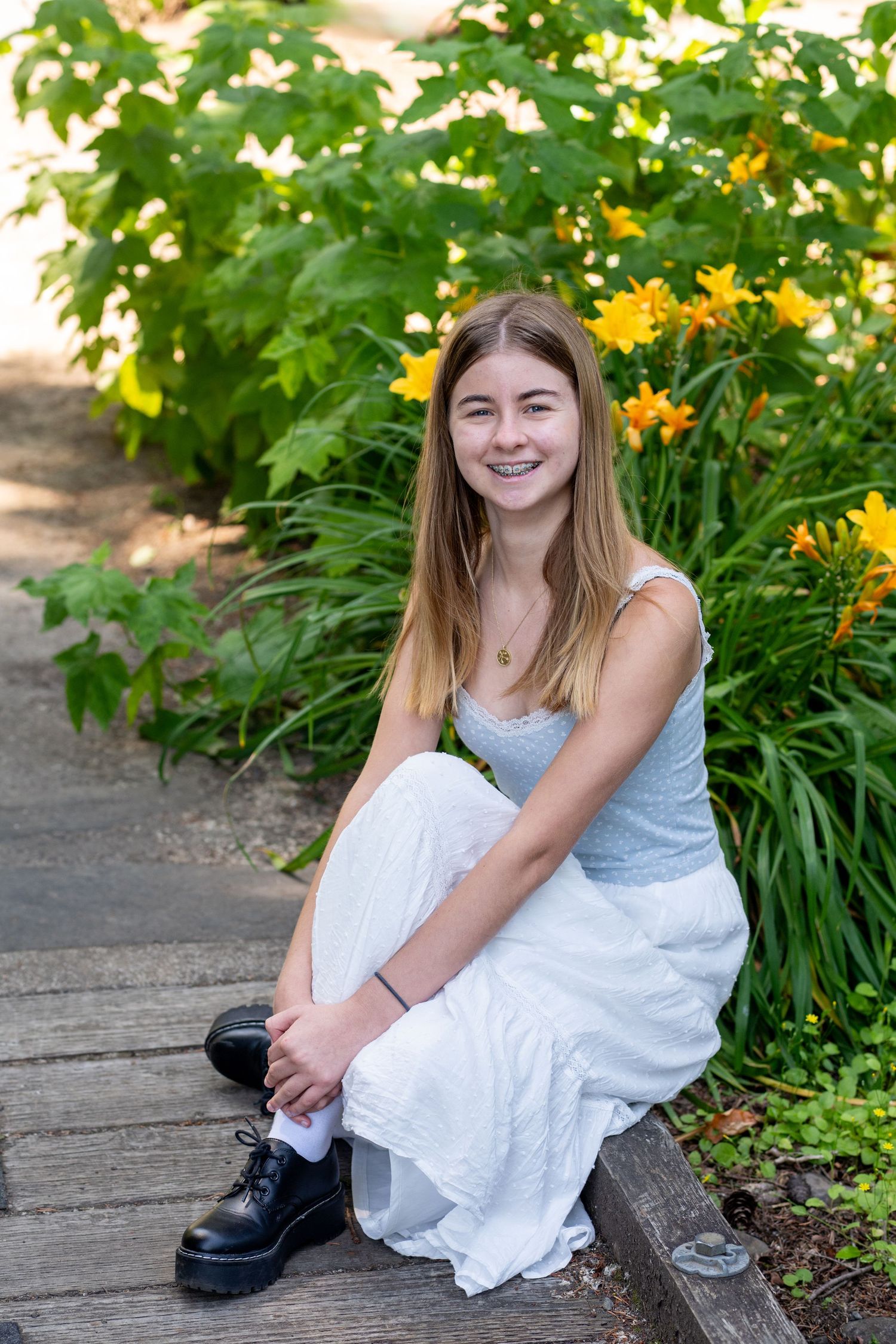 A person in a white dress sits smiling beside a garden of yellow flowers and greenery.