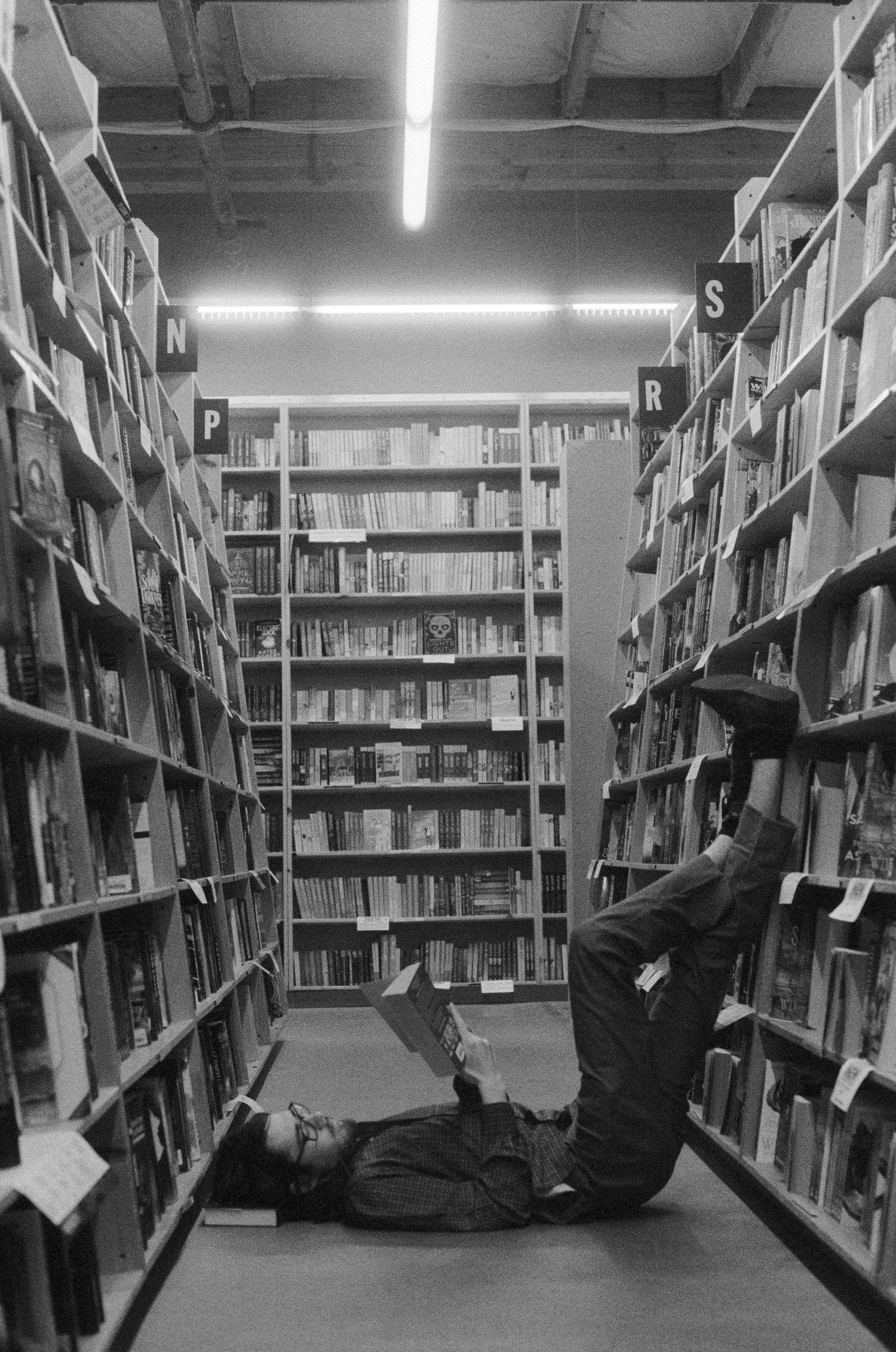 Black and white photo of people relaxing between tall library bookshelves filled with books.