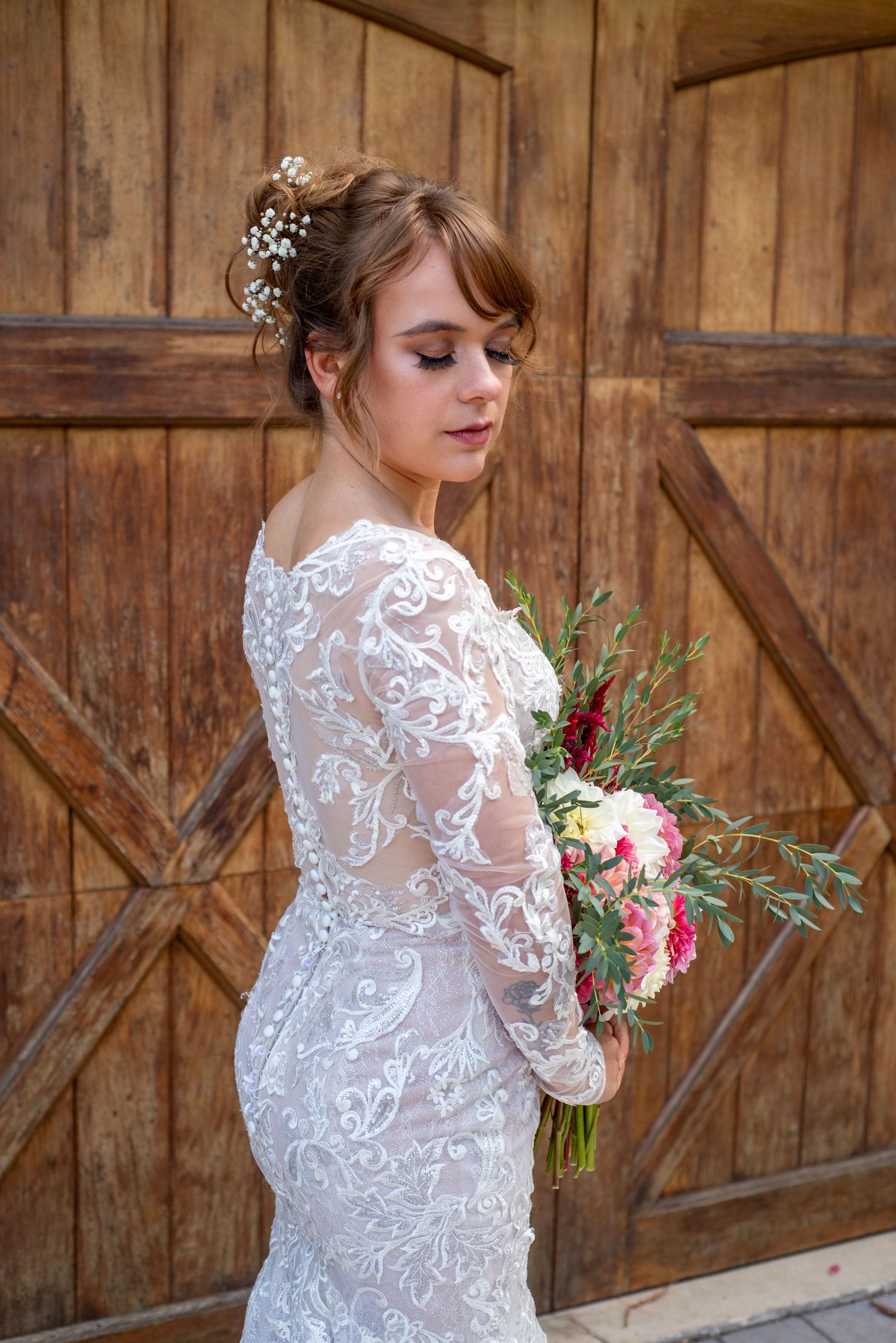 A bride in a lace wedding dress with long sleeves holds a bouquet against a rustic barn door.