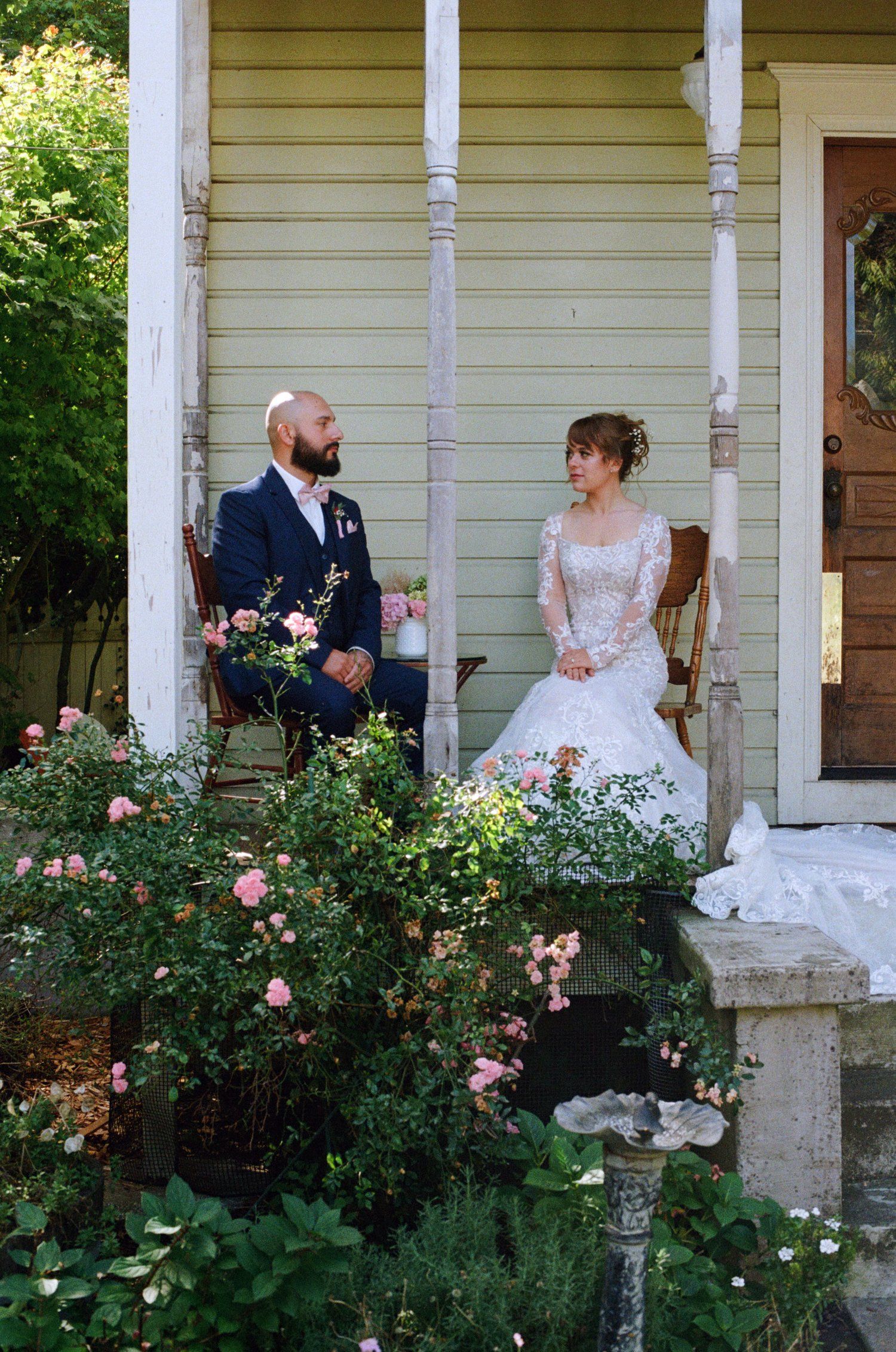 A couple sits on a rustic porch surrounded by blooming rose bushes on their wedding day.