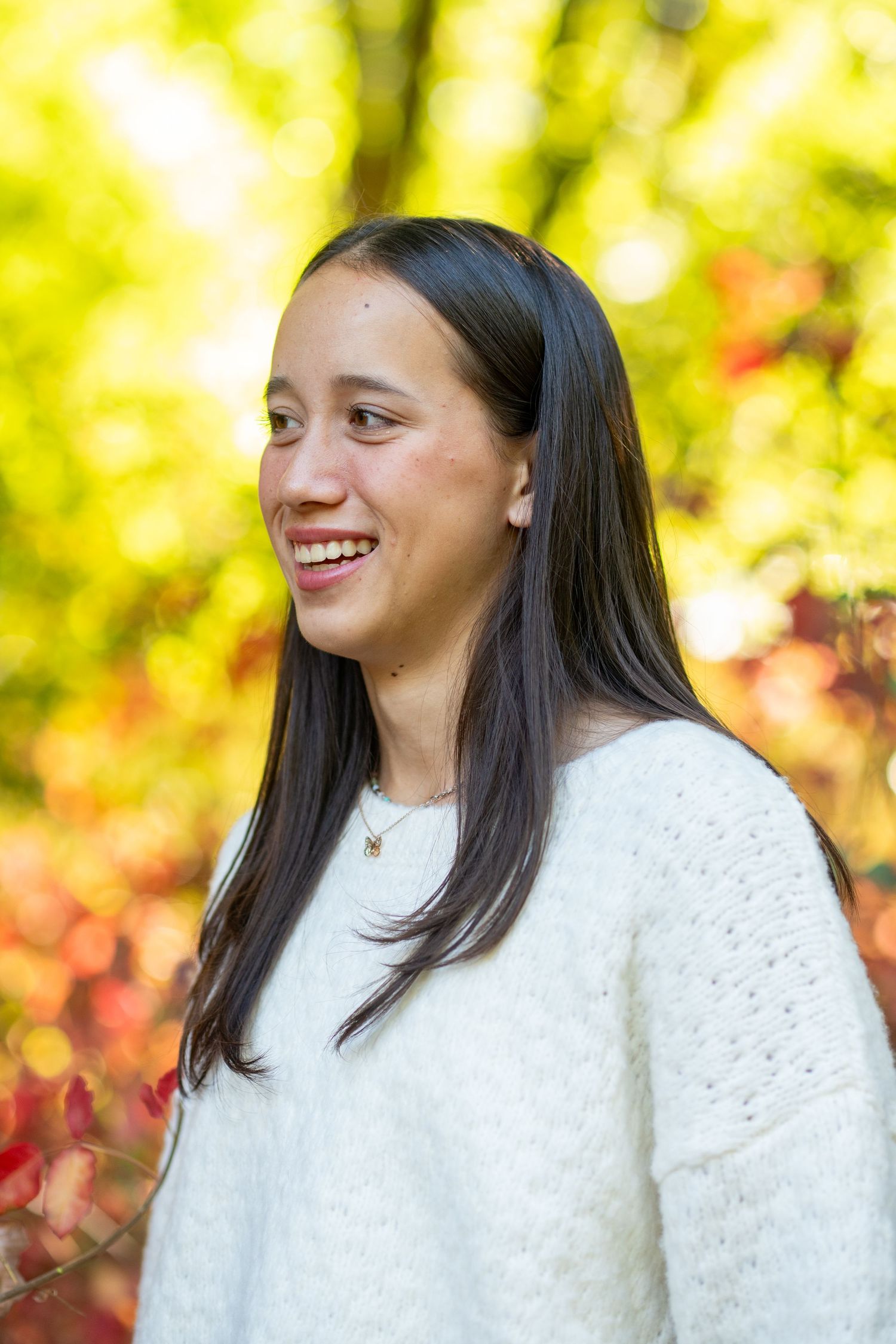 Person wearing a white sweater smiles brightly against autumn foliage in an outdoor portrait session.