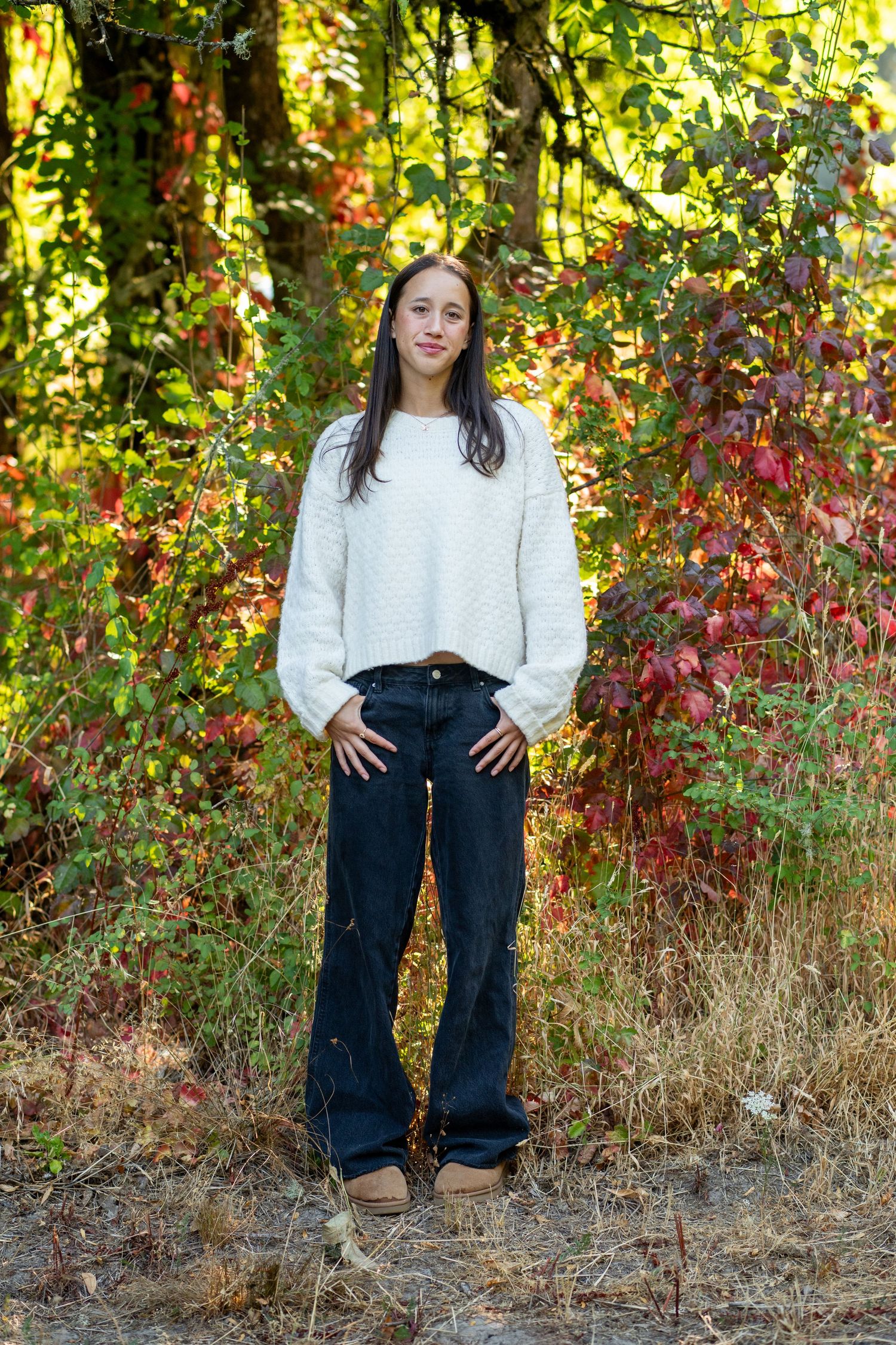 A white sweater and dark jeans outfit is showcased against a backdrop of vibrant autumn foliage.