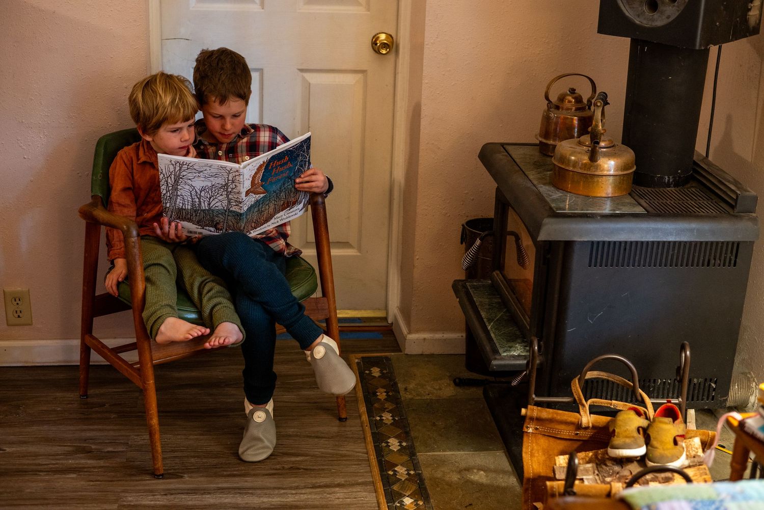 A cozy reading scene by a wood stove with a book being shared in a wooden chair, with teapots nearby.