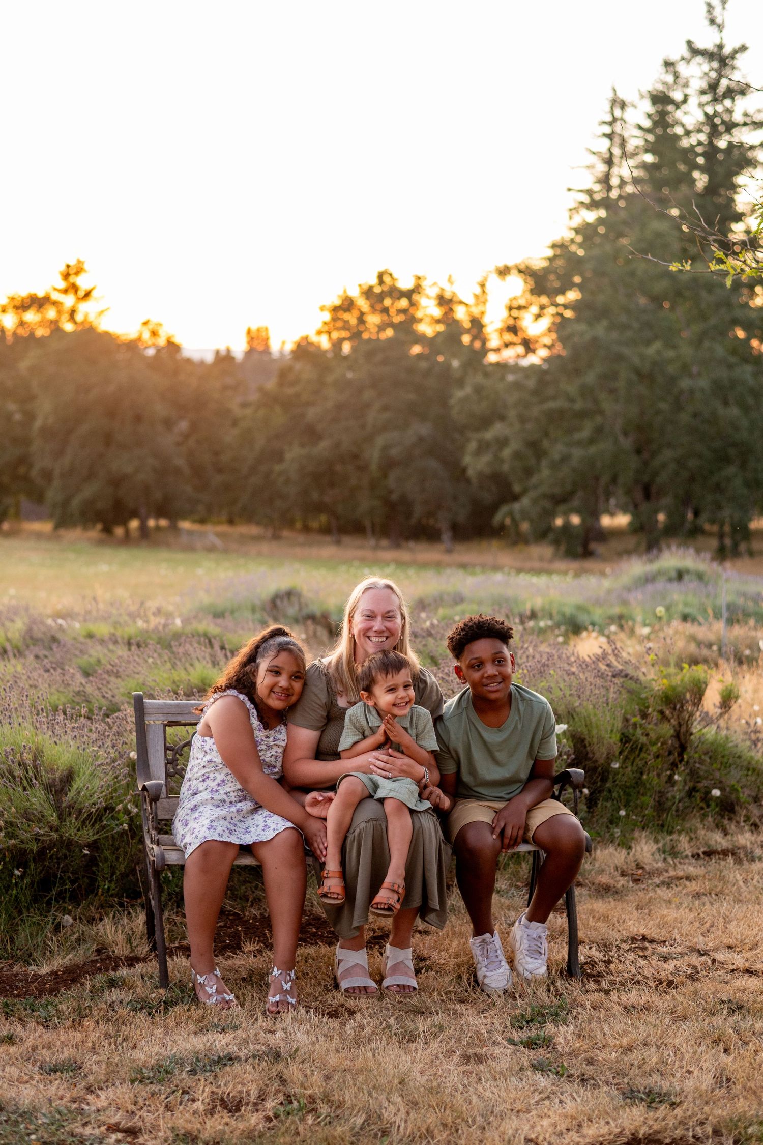 A group sits together on a rustic bench in a field at sunset with evergreen trees in the background.