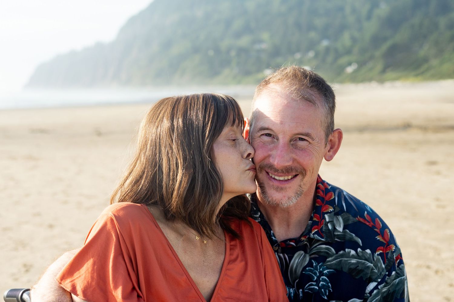 A couple shares tender moments together on a sunny beach while wearing casual attire and smiling together.