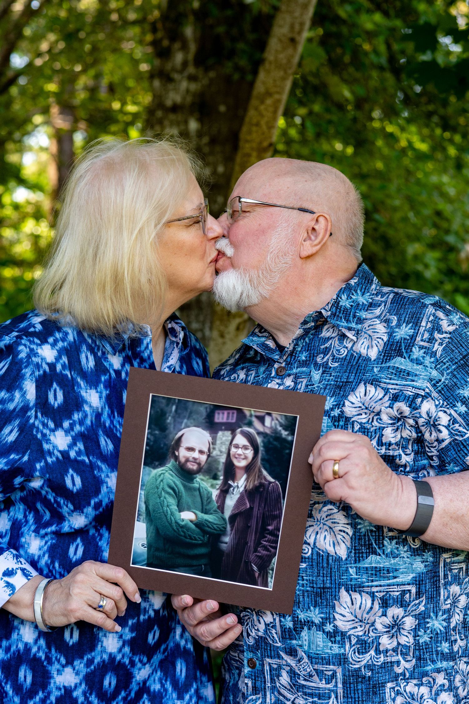 A couple in matching blue Hawaiian shirts share affectionate moments while holding and displaying a framed photograph.