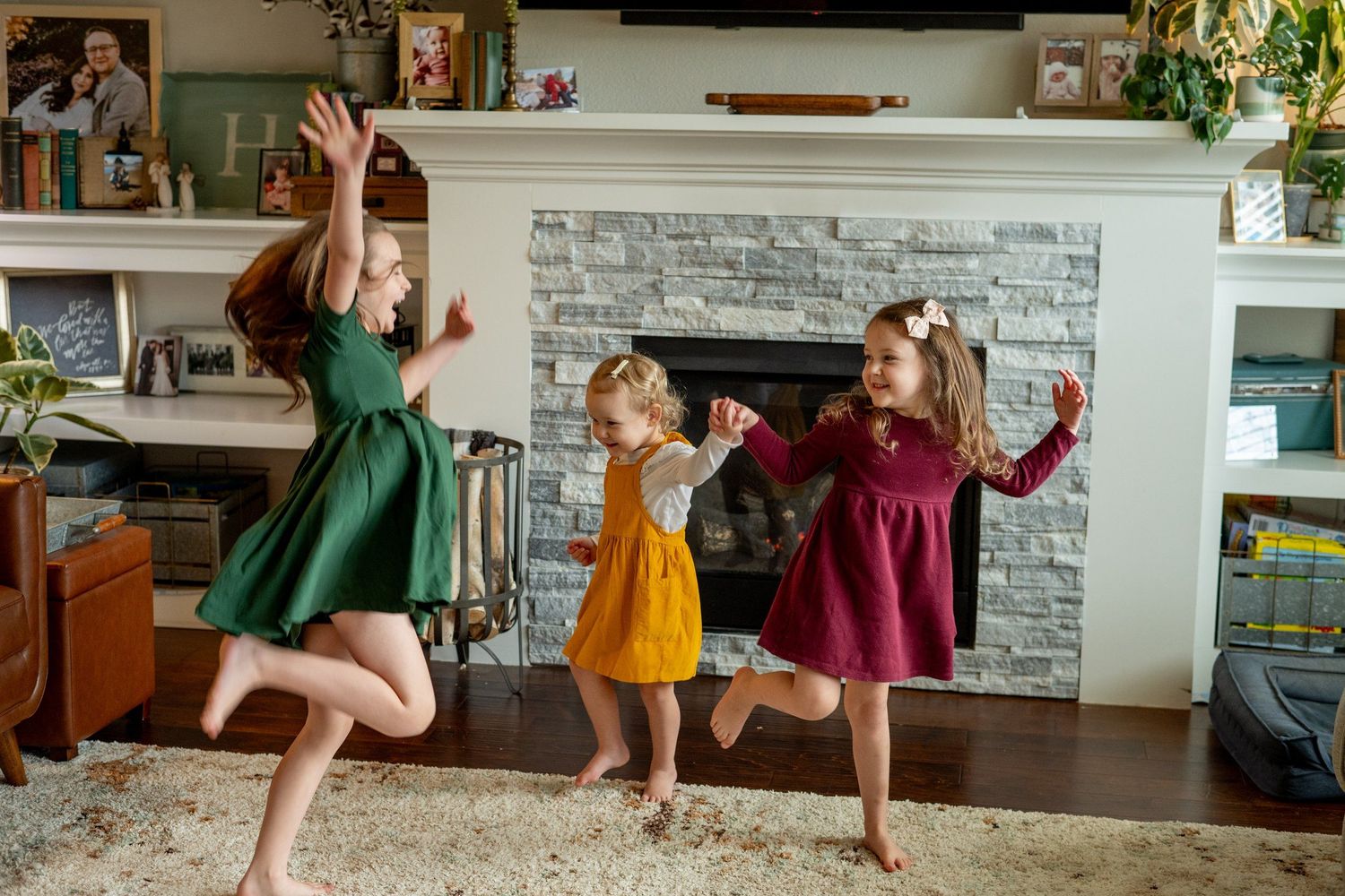 Three children in colorful dresses dancing and jumping together in a living room.