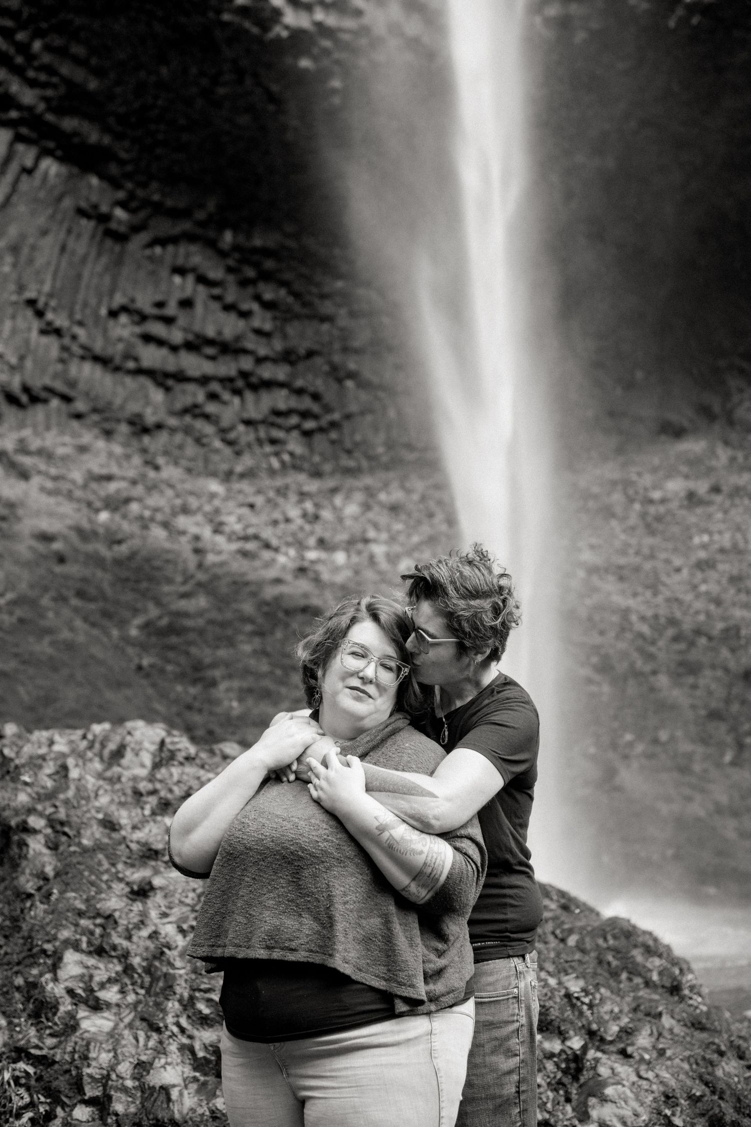 A couple shares an intimate moment together in front of a cascading waterfall in a scenic mountainous landscape.