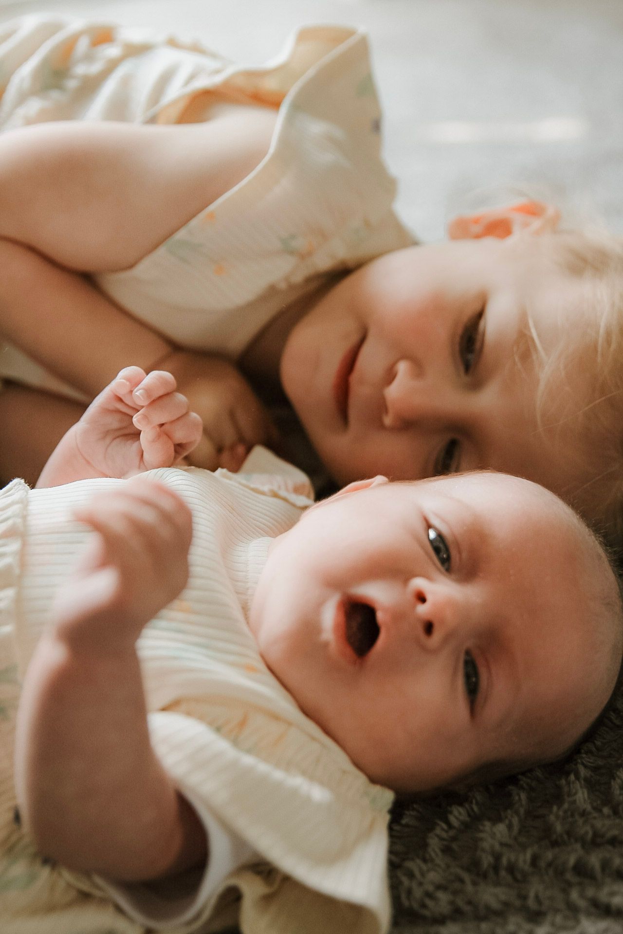 A smiling toddler girl lies beside her laughing newborn baby sibling on a soft grey rug.