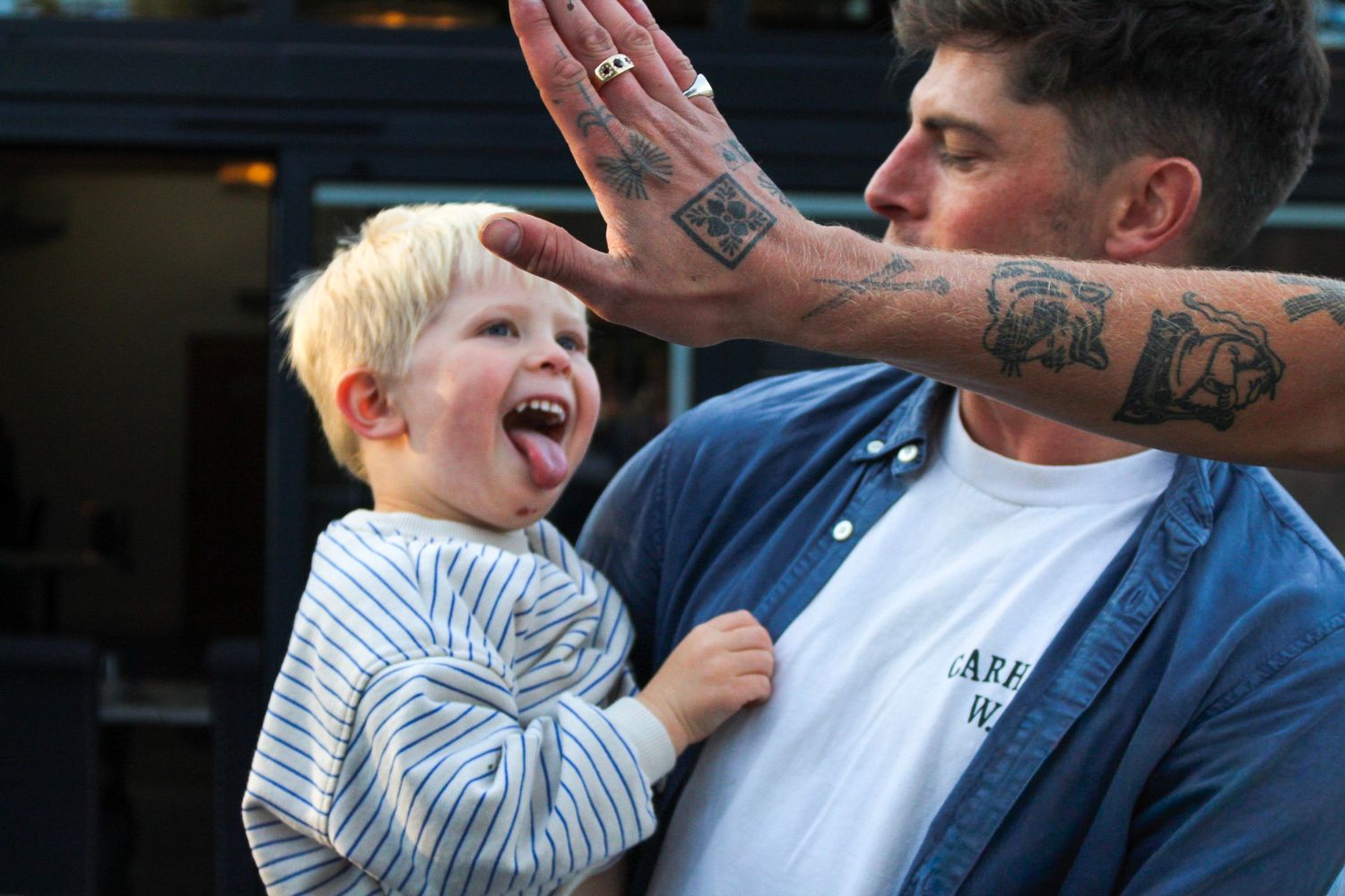 Tattooed father laughing with toddler son giving a high five outdoors.