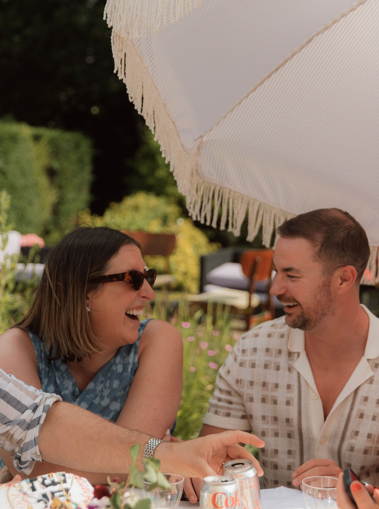 A couple laughing together at an outdoor garden table beneath a fringed white umbrella on a sunny day.