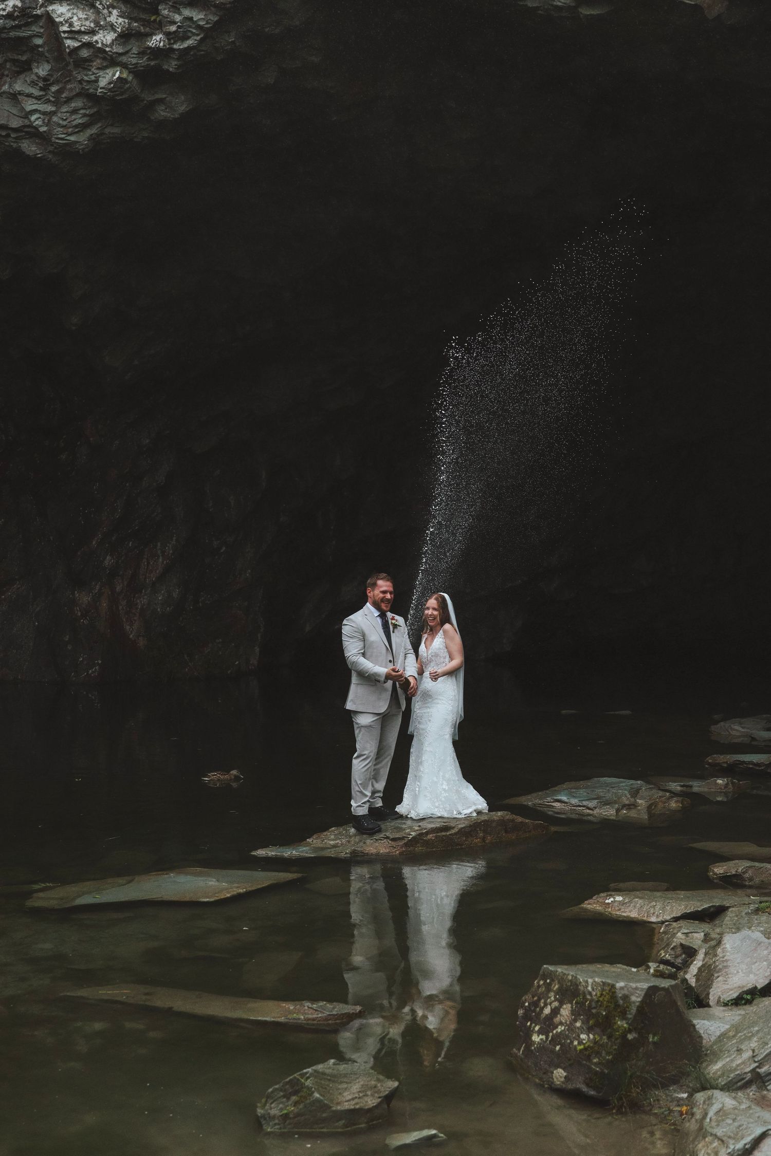 Bride and groom stand on stepping stones in a dark cave pool, spraying champagne into the air during their wedding photoshoot.