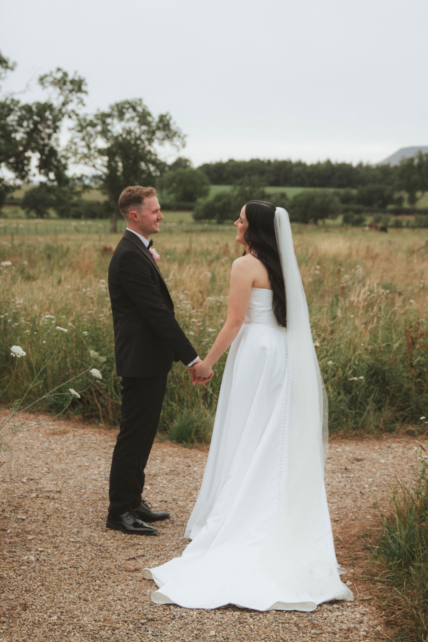 Bride in a white gown and long veil holds hands with a smiling groom in a dark suit on a gravel path beside a wildflower meadow.