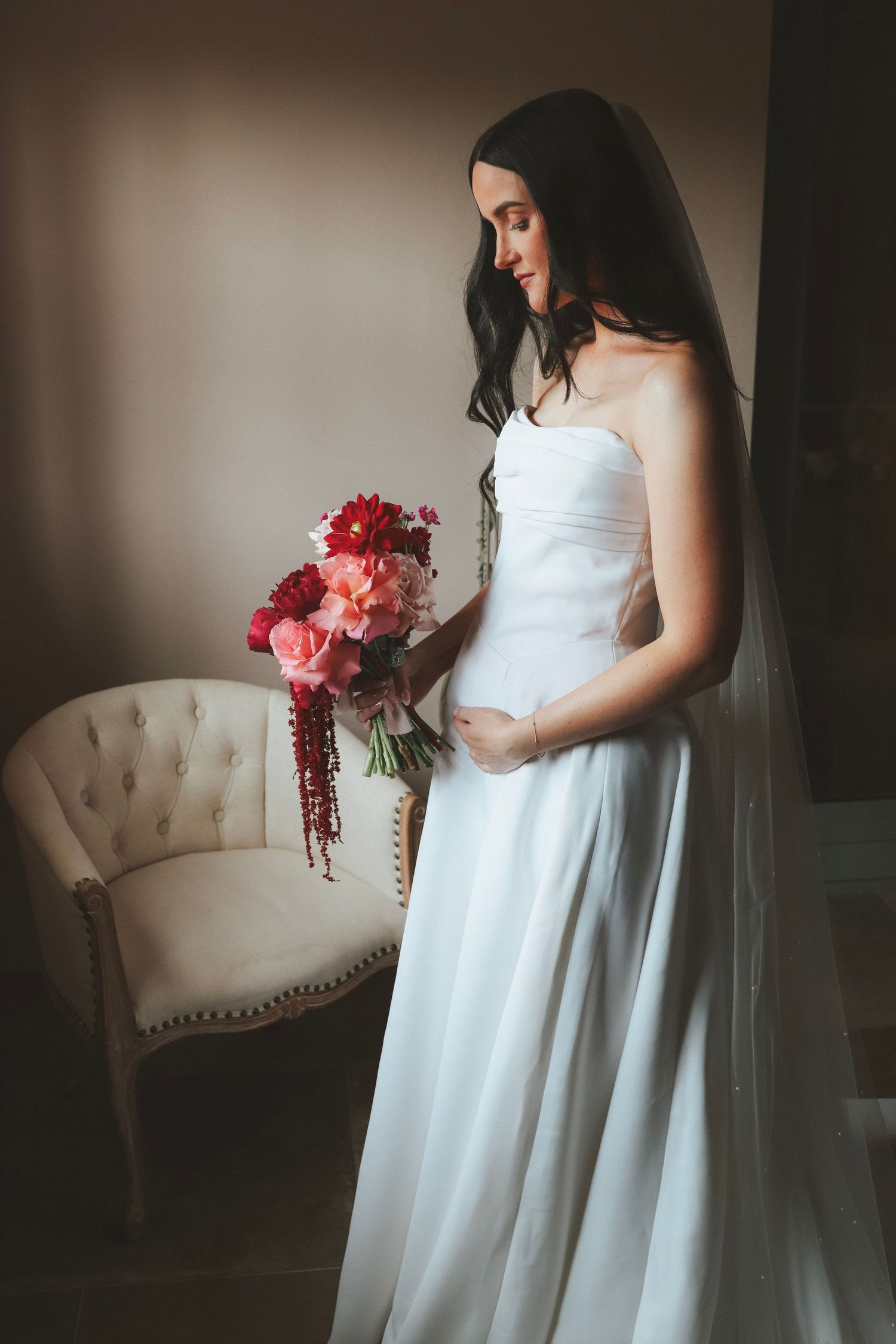Bride in a strapless white gown and long veil holds a red and pink cascading bouquet beside a tufted cream chair in soft window light.