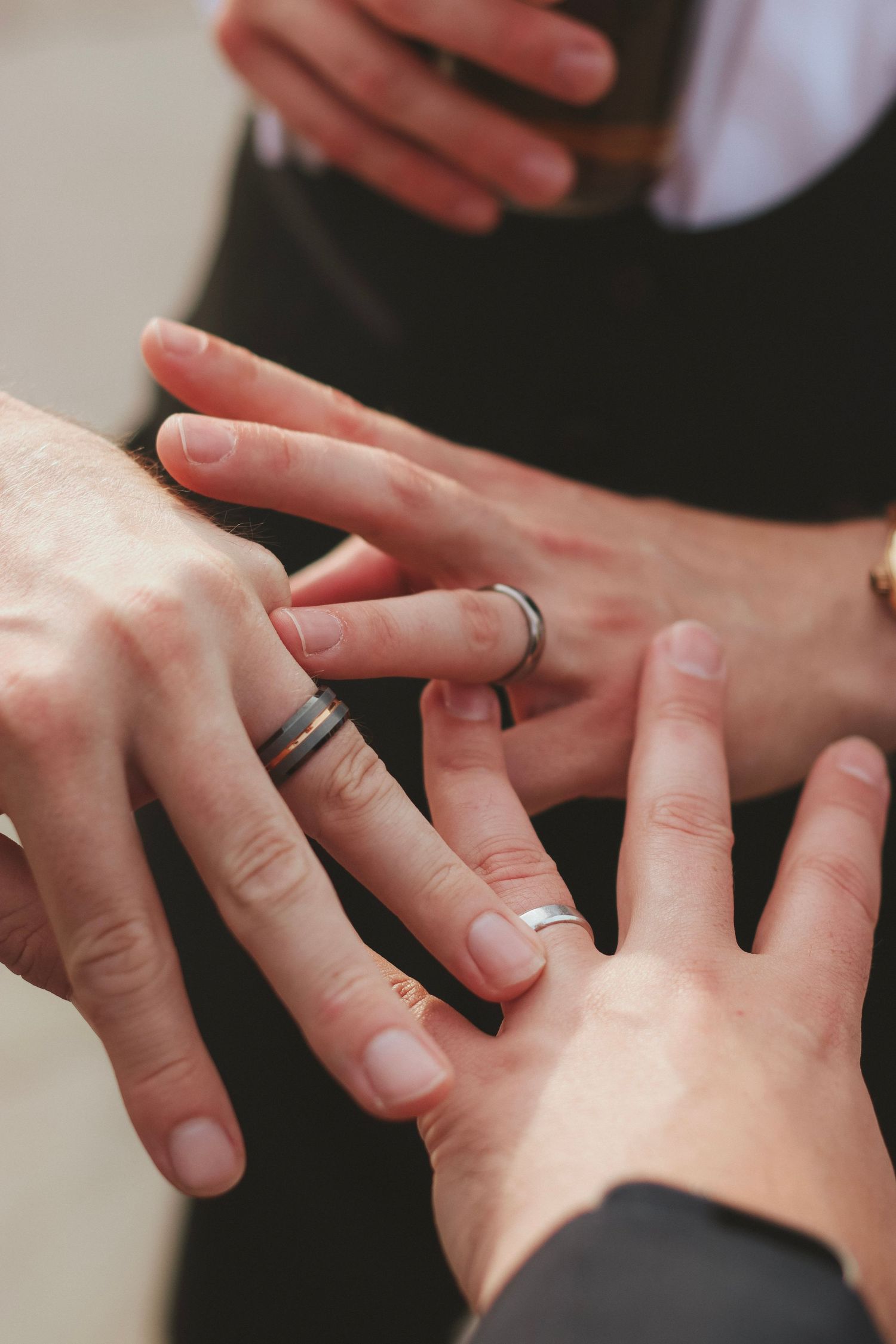 Close-up of several hands comparing and exchanging silver wedding rings during a ceremony, with a man in a dark suit in the background.