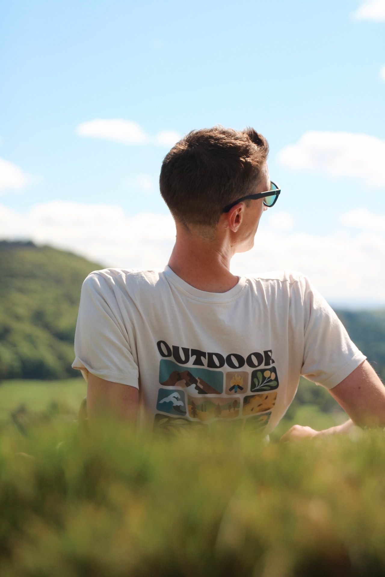 Young man in an Outdoor graphic t-shirt and sunglasses sitting on a hillside with scenic green valley views.