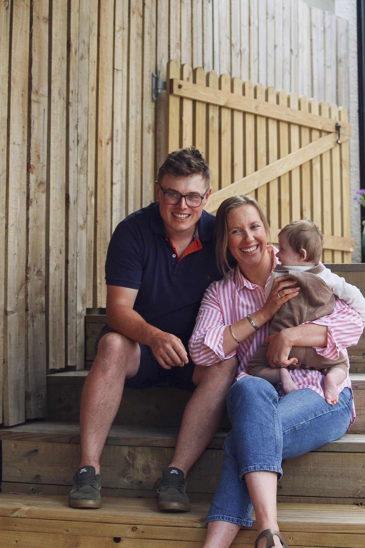 A smiling young family with a baby sits together on wooden steps outside their home.