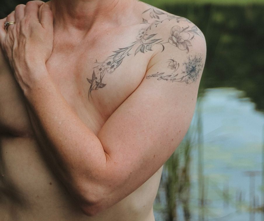 A person with floral tattoos on their shoulder and chest crosses their arms near a body of water.
