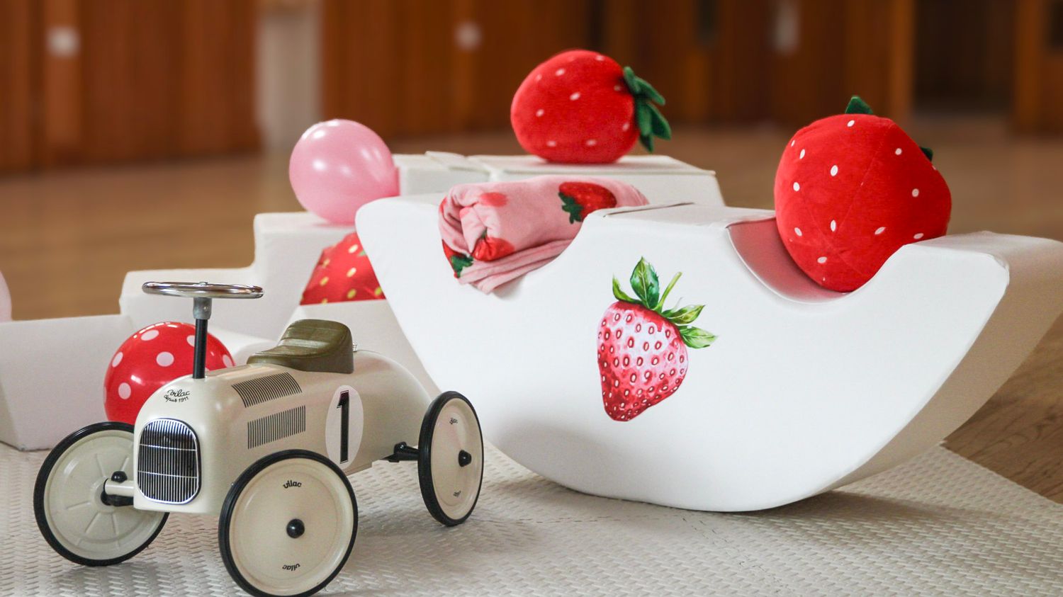Strawberry-themed party decorations with white rocking toys, balloons and oversized strawberry props on a wooden floor.