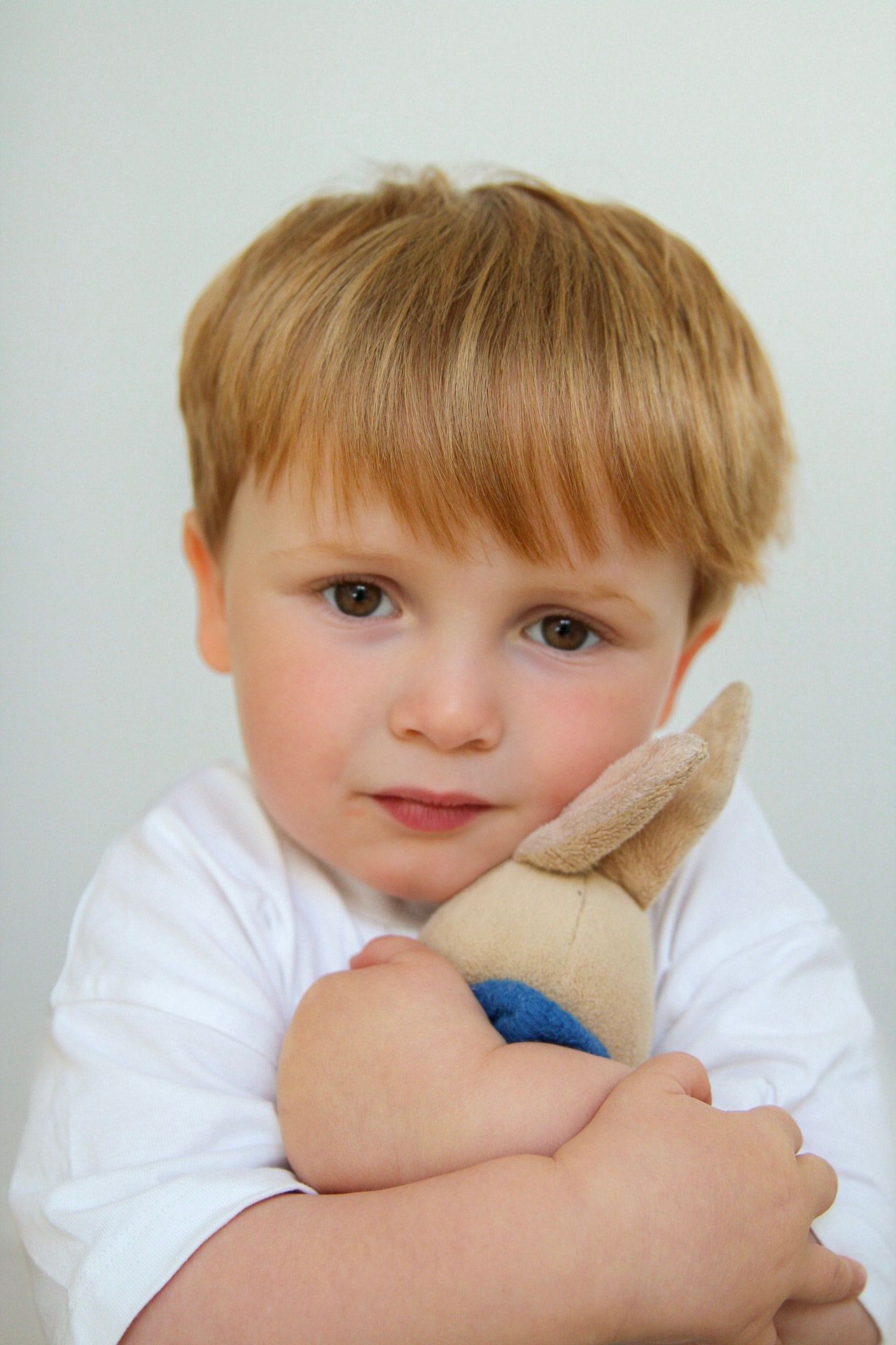Cute blonde toddler boy in white shirt hugging small stuffed animal toy against white background.