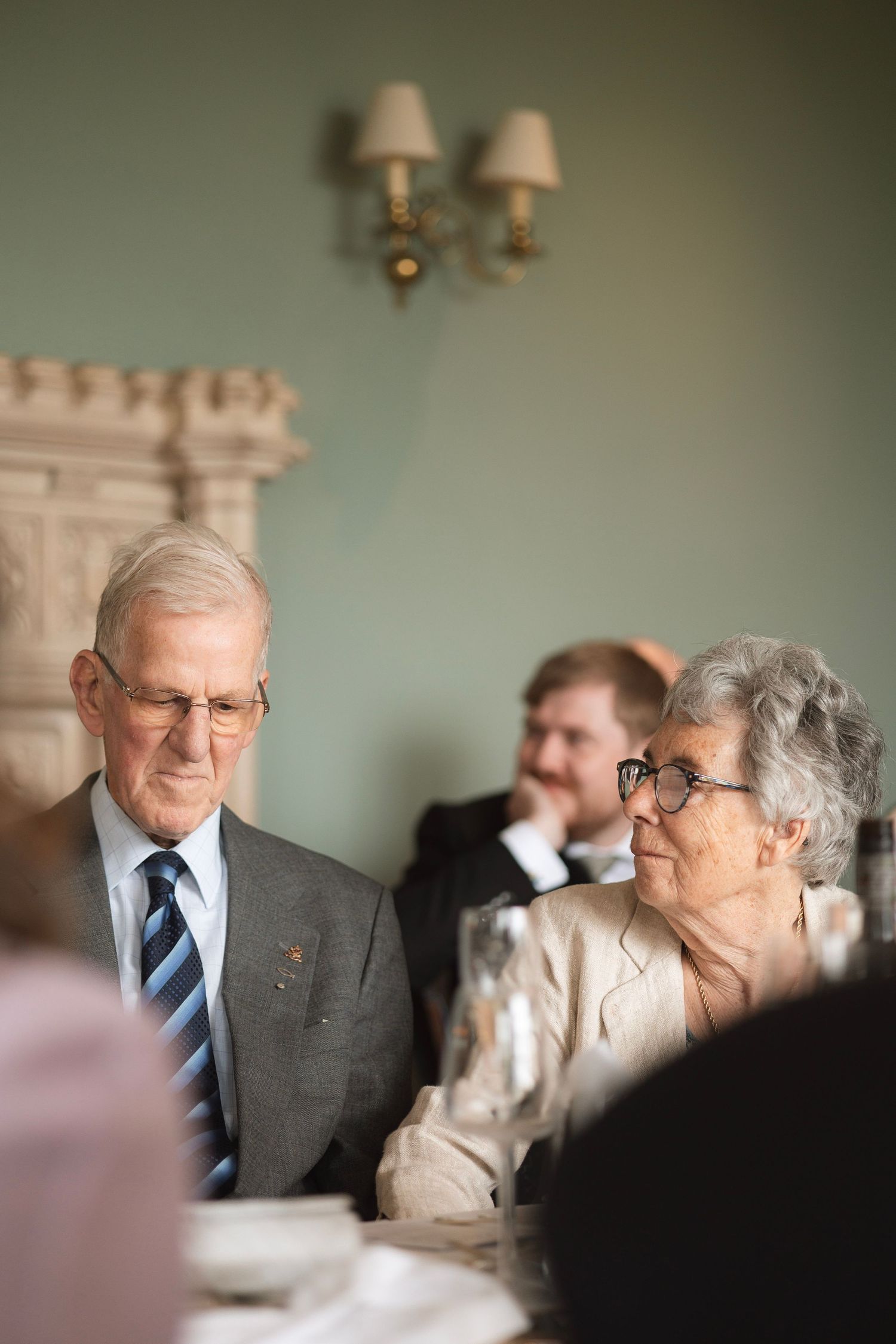 Elderly couple in formal attire seated at a dining table during an elegant event, man in suit and tie, woman in glasses.