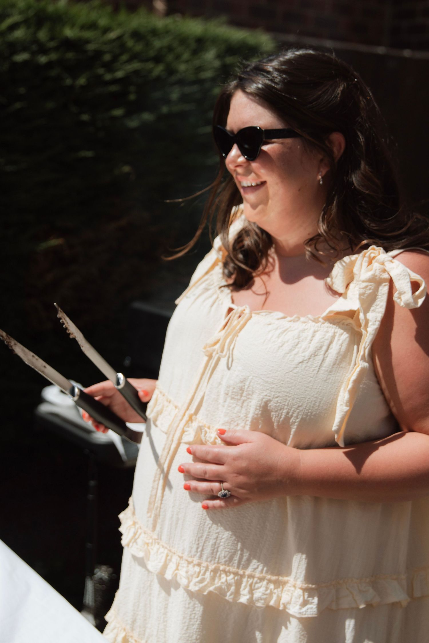 A smiling pregnant woman in a cream ruffled dress and sunglasses holds scissors at an outdoor event.