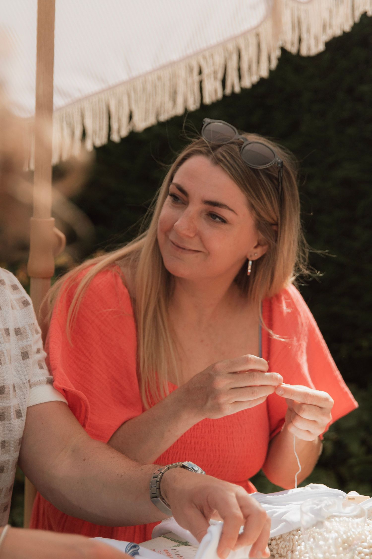 Smiling woman in a coral top with sunglasses on her head sits outdoors under a fringed parasol, holding a delicate bead strand.