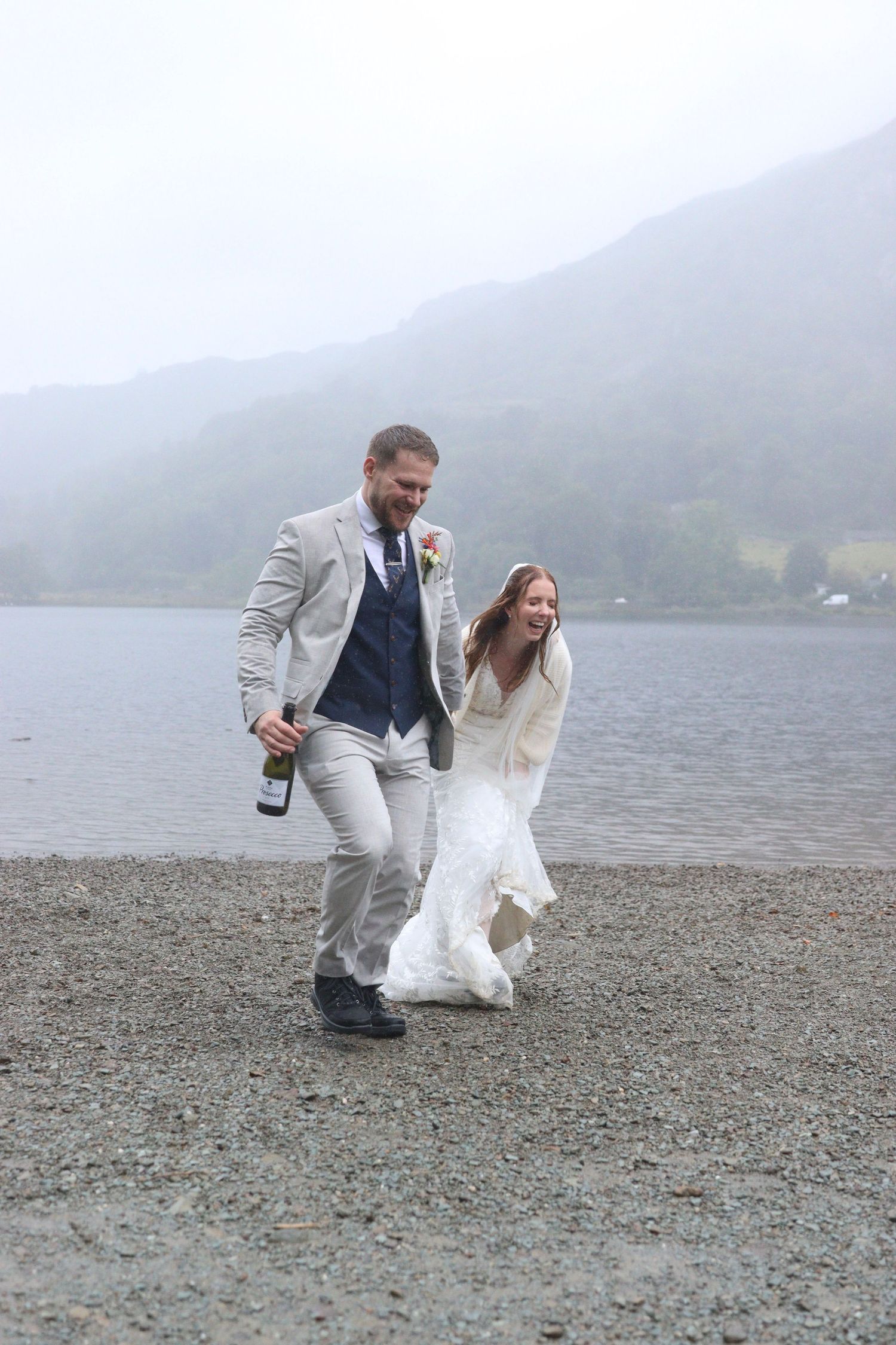 Laughing bride in a white lace gown and groom in a grey suit holding a bottle run along a misty lakeshore on their wedding day.