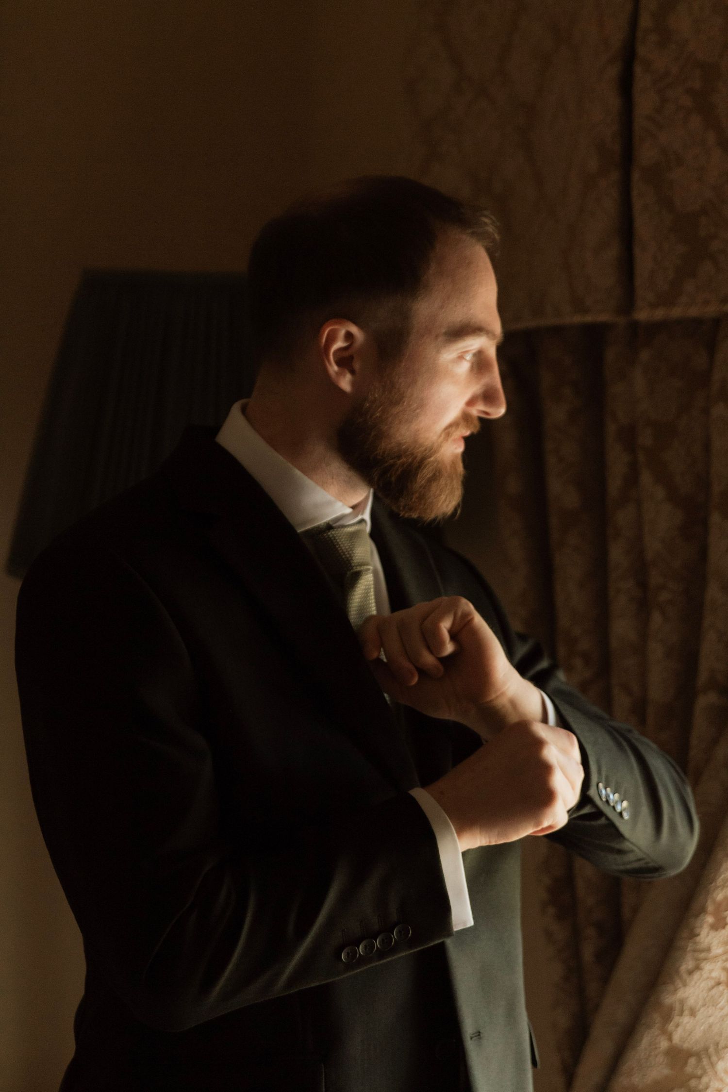 Bearded groom in black suit adjusting cufflinks by window in dramatic moody lighting.