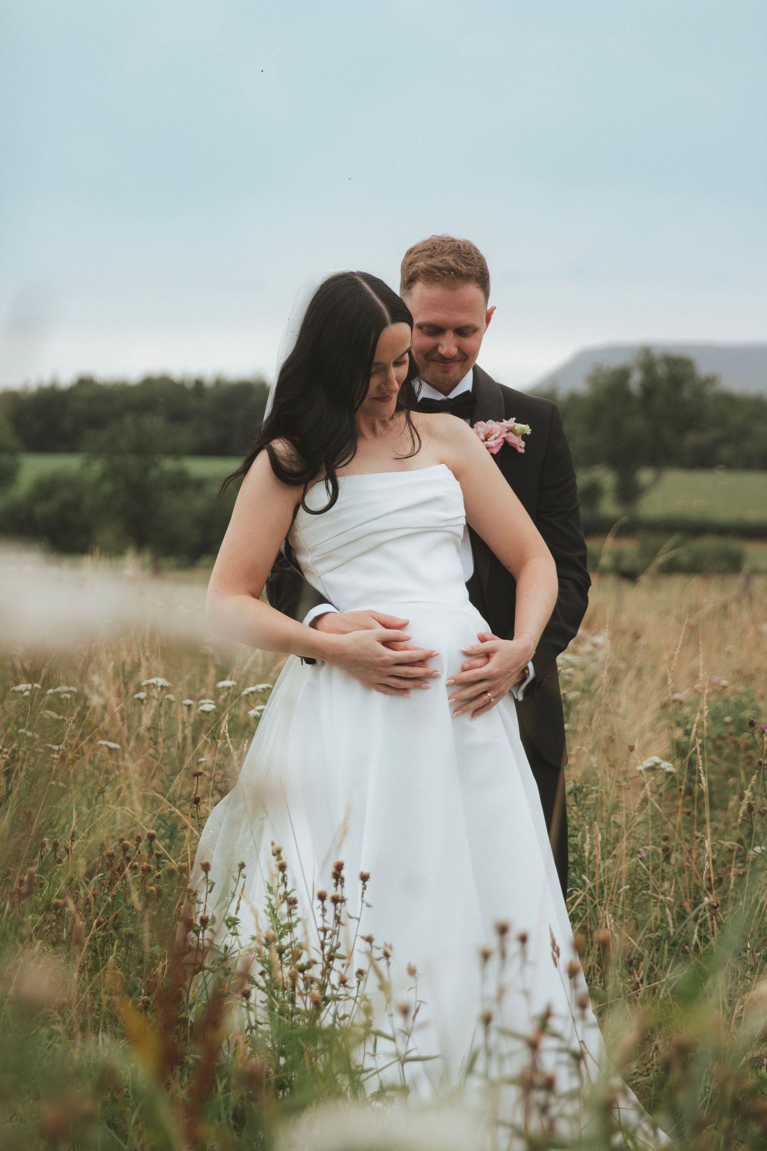 Bride in a white strapless gown and groom in a black suit embrace in a wildflower meadow, his hands resting on her waist.