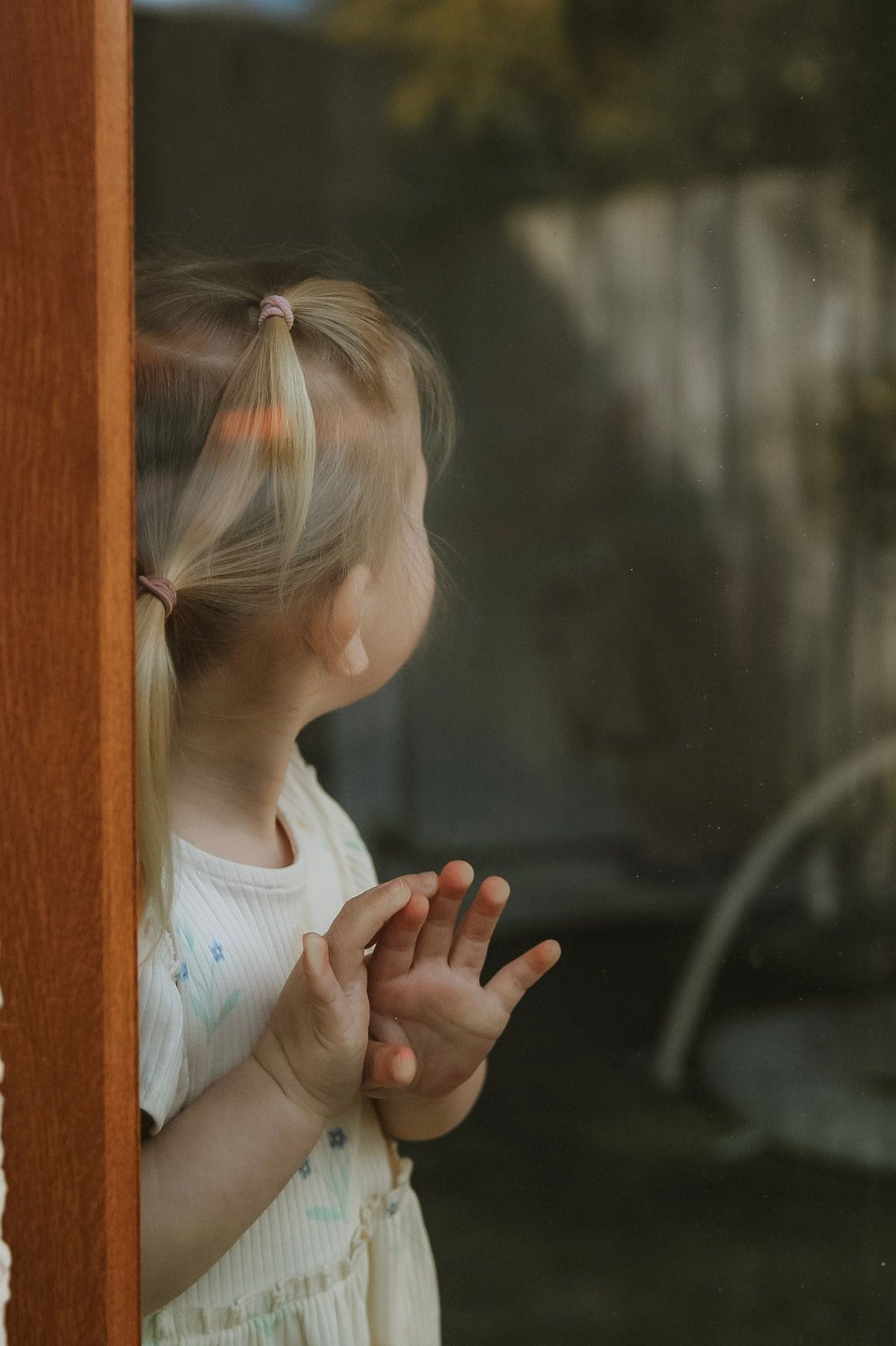 Young blonde girl in white dress pressing hands against glass door looking outside.