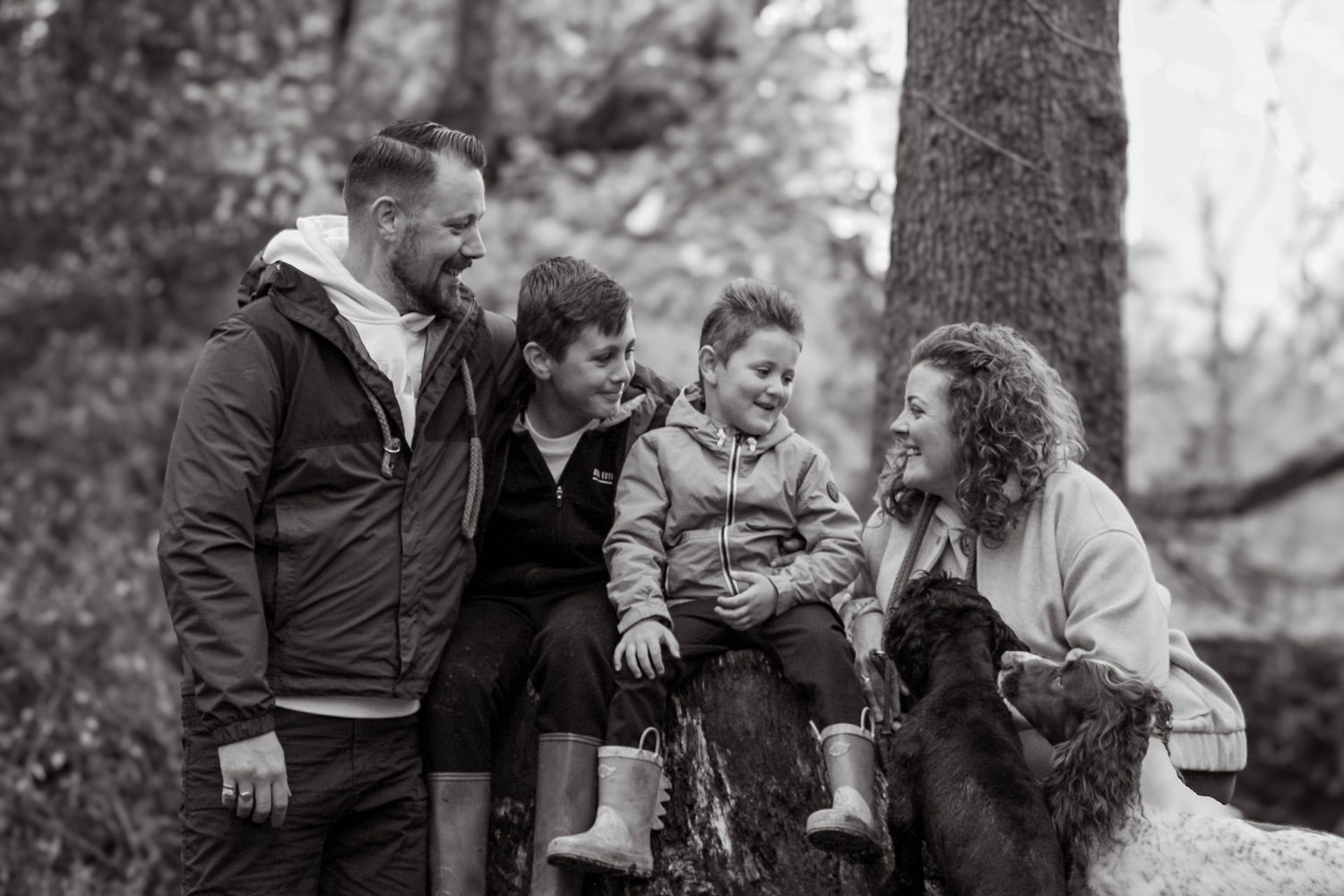 A happy family of four with two children smiles together in a black and white forest photo.