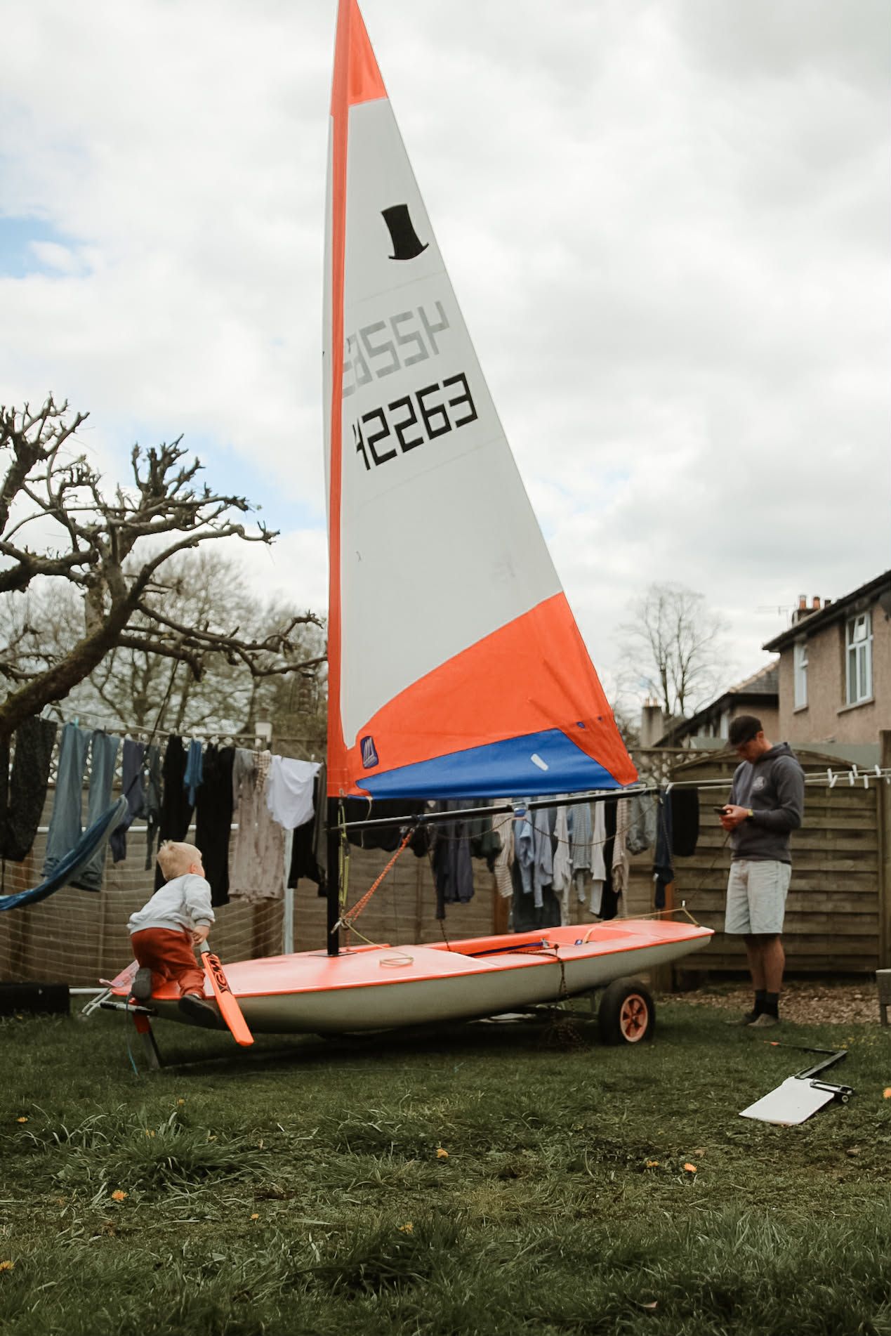 Child and adult examining a small orange and white sailboat with number 42263 in a backyard.