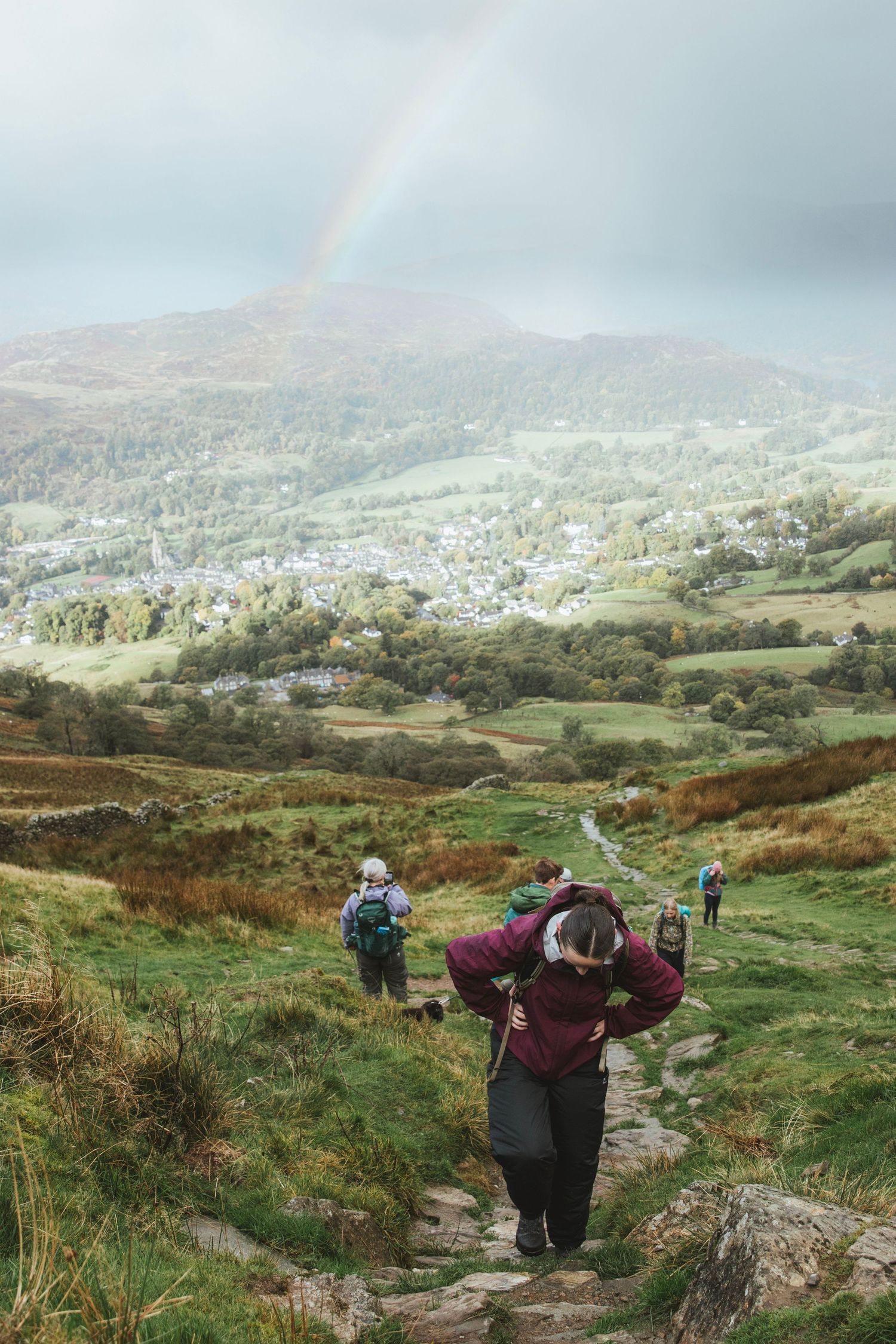 Hikers climb a rocky mountain path with a rainbow arching over green hills and a small village in the valley below.