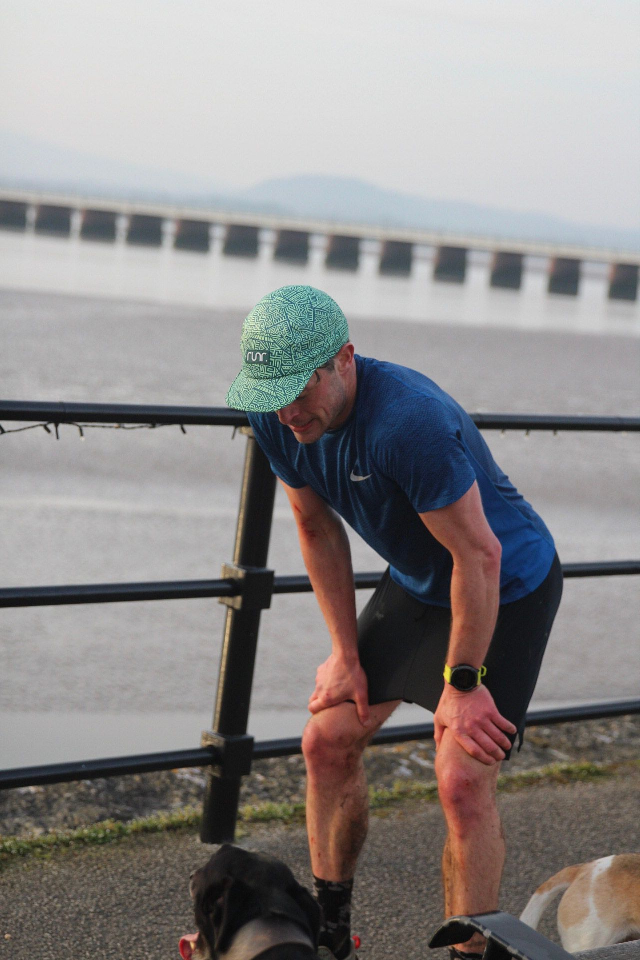 A male runner in a blue Nike shirt and green cap leans on a railing to rest by the waterfront.