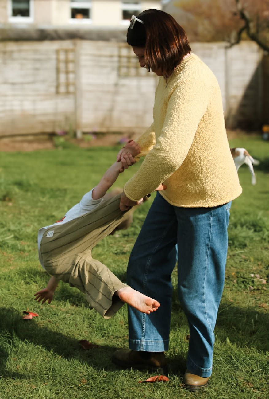 Mother in yellow sweater and jeans playfully swinging her barefoot child in a sunny backyard.