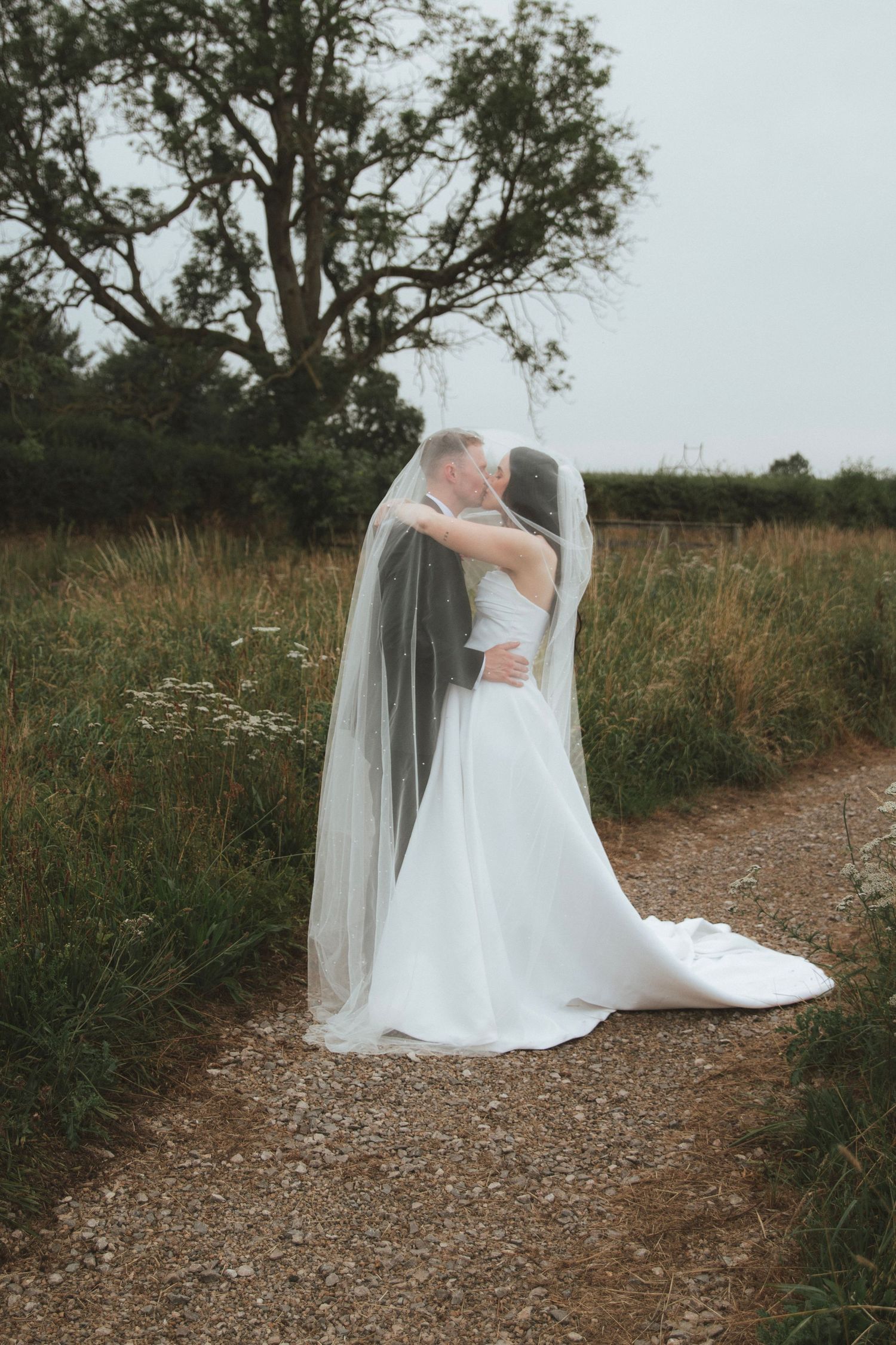 Bride and groom share a kiss under a flowing veil on a gravel country path, surrounded by tall grass and trees on a overcast day.
