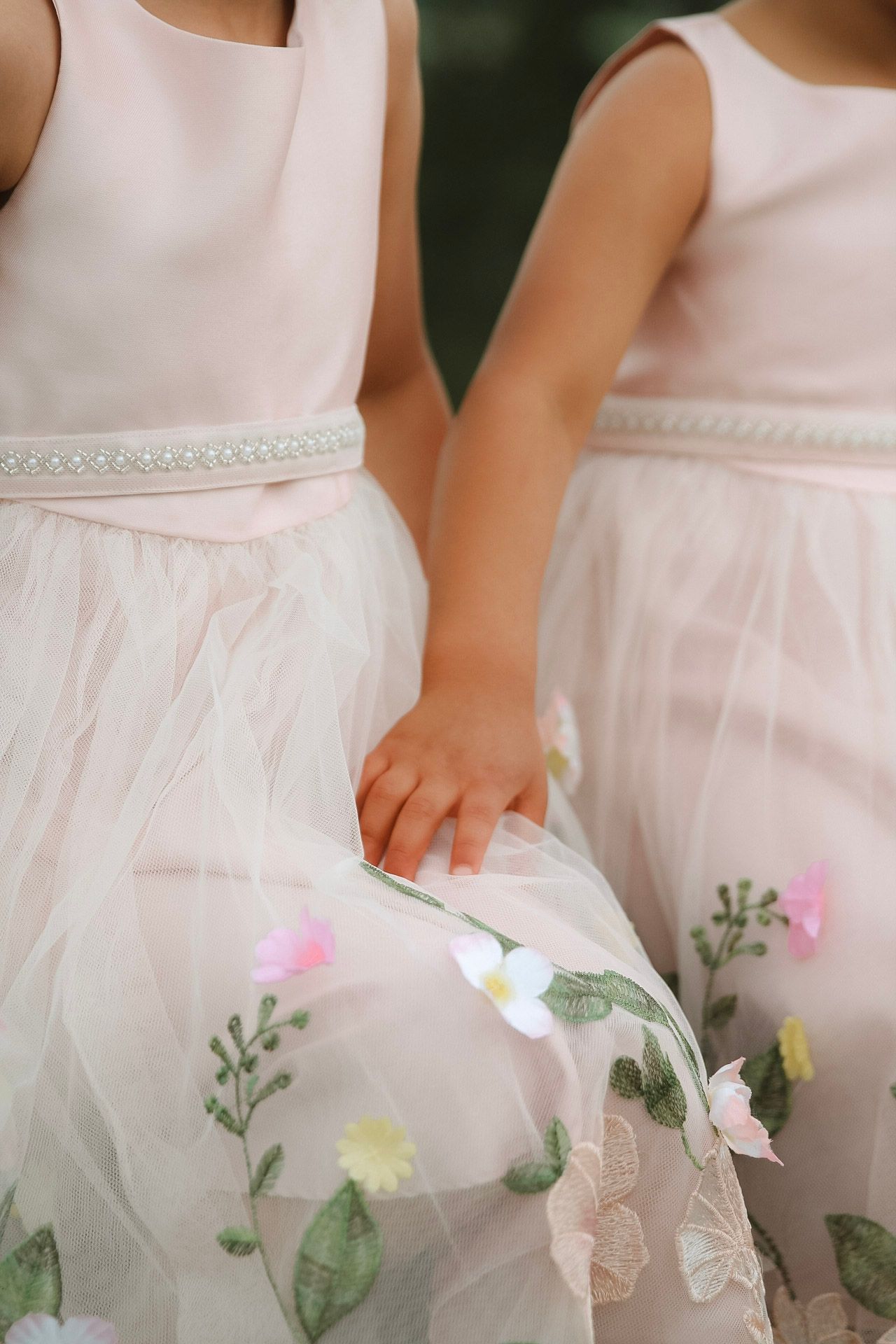 Two young flower girls in blush pink tulle dresses with floral embroidery and pearl sash belts holding hands.
