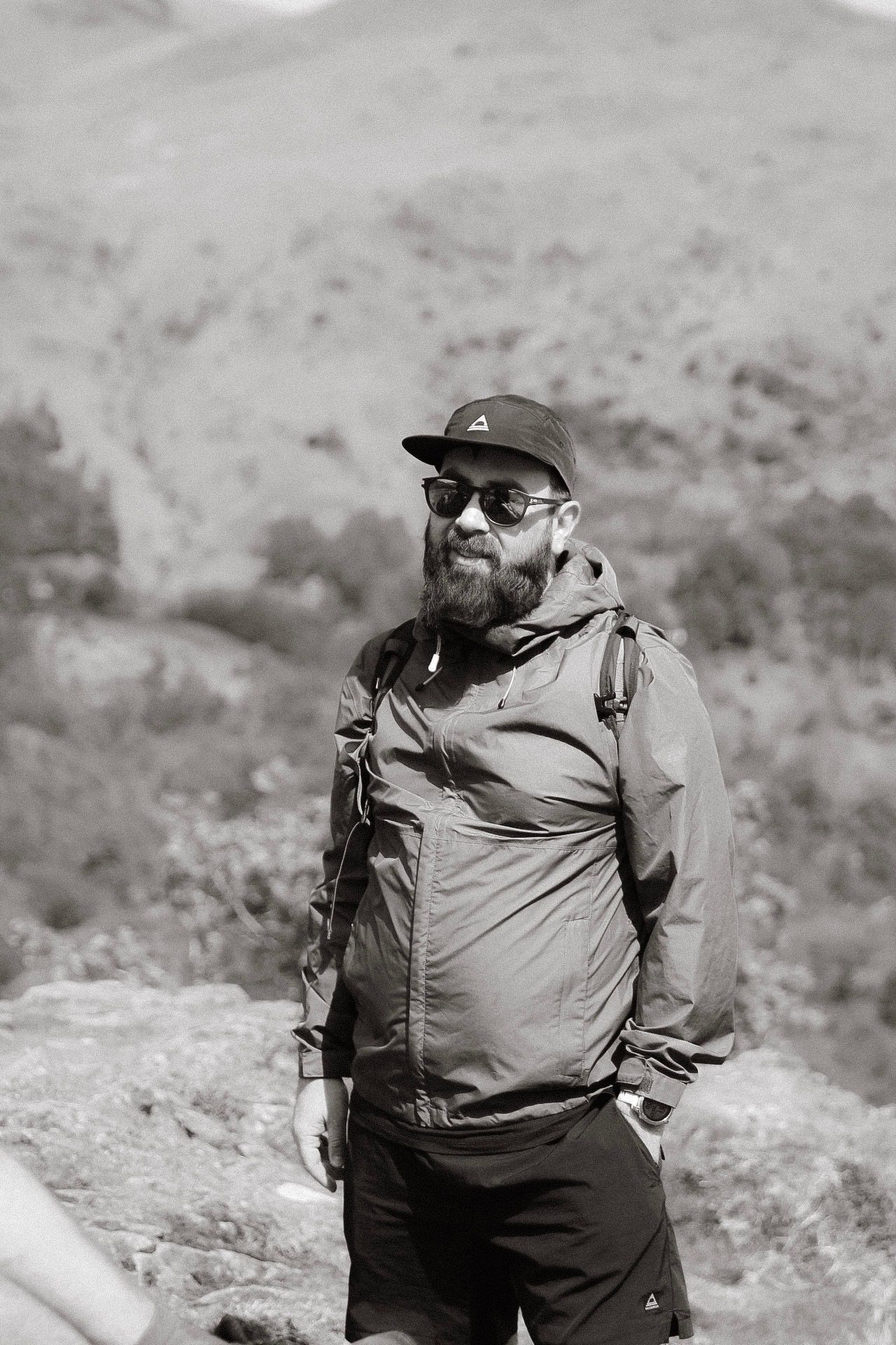 Bearded man in cap, sunglasses and hiking backpack standing in a rocky mountain landscape, black and white photo.